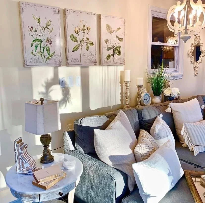 Living room with beige walls, three botanical art prints, a gray sofa with white and beige pillows, a round white side table with a lamp, candles, and decorative items, a window with a white frame, and a chandelier overhead.
