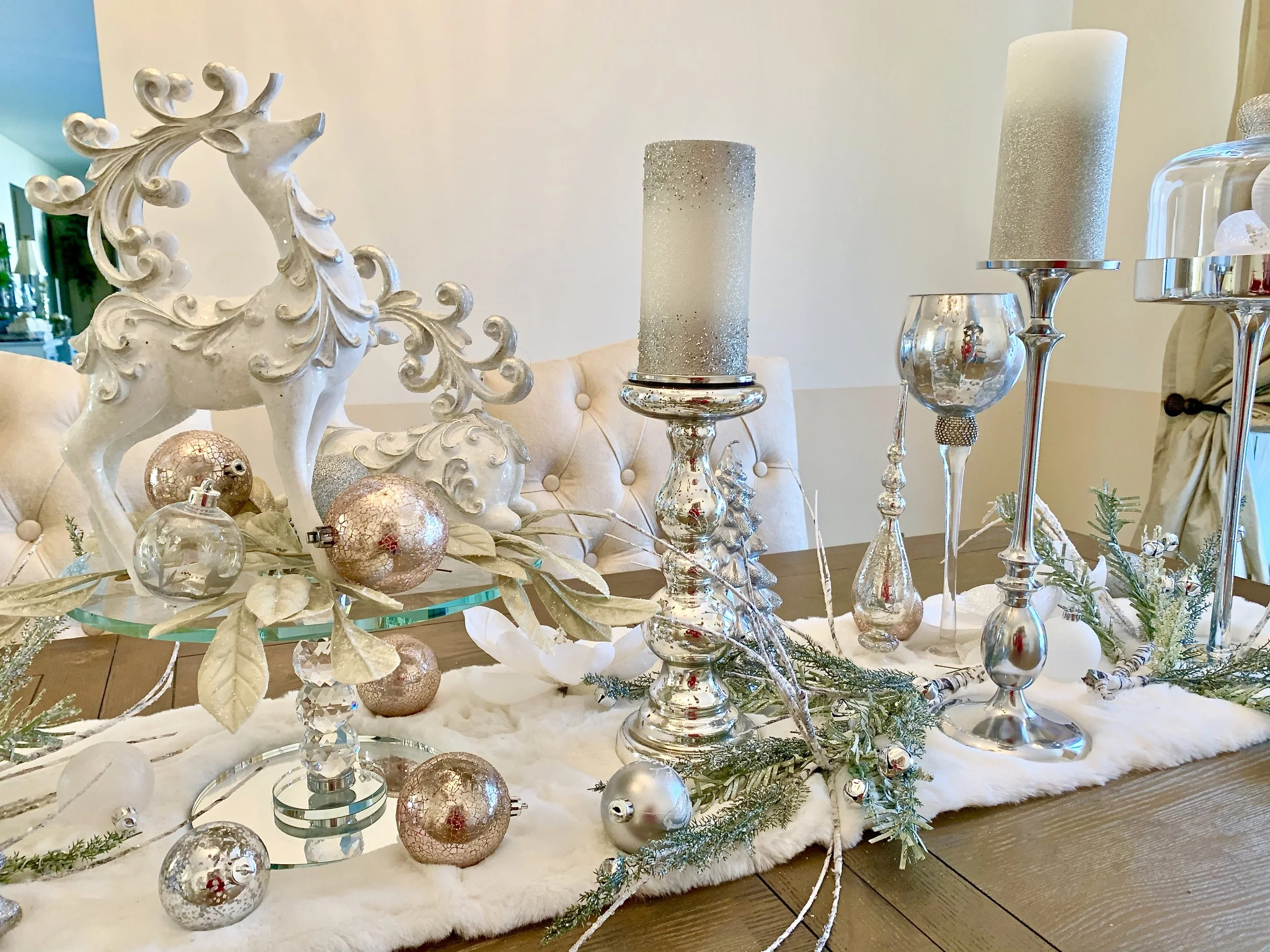 Decorative Christmas table centerpiece with silver and white ornaments, candles, greenery, and a white reindeer figurine on a dining table.
