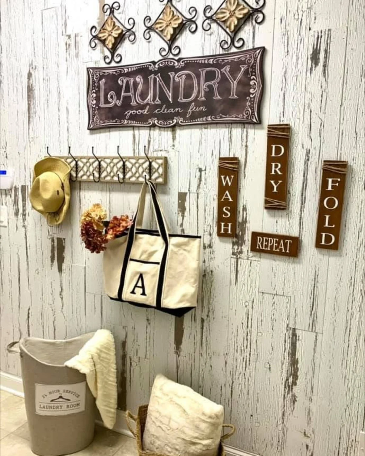 A laundry room wall decorated with signs and storage items. There is a large sign that says 'LAUNDRY good clean fun', three brown signs that say 'WASH', 'DRY', and 'FOLD', and a small sign that says 'REPEAT'. A hook rack with a beige hat, a floral dr