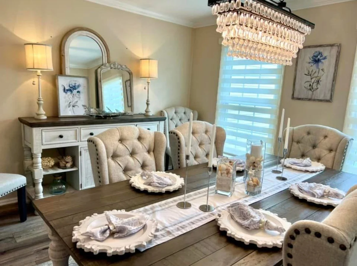 A dining room with a wooden table, beige tufted chairs, a crystal chandelier, a sideboard with lamps, and decorative plates set for a meal.
