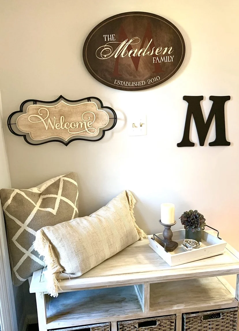 Decorative wall with signs reading 'The Madsen Family,' 'Welcome,' and a large black letter 'M,' above a wooden bench with throw pillows and a tray holding a candle, a decorative plant, and other small items.