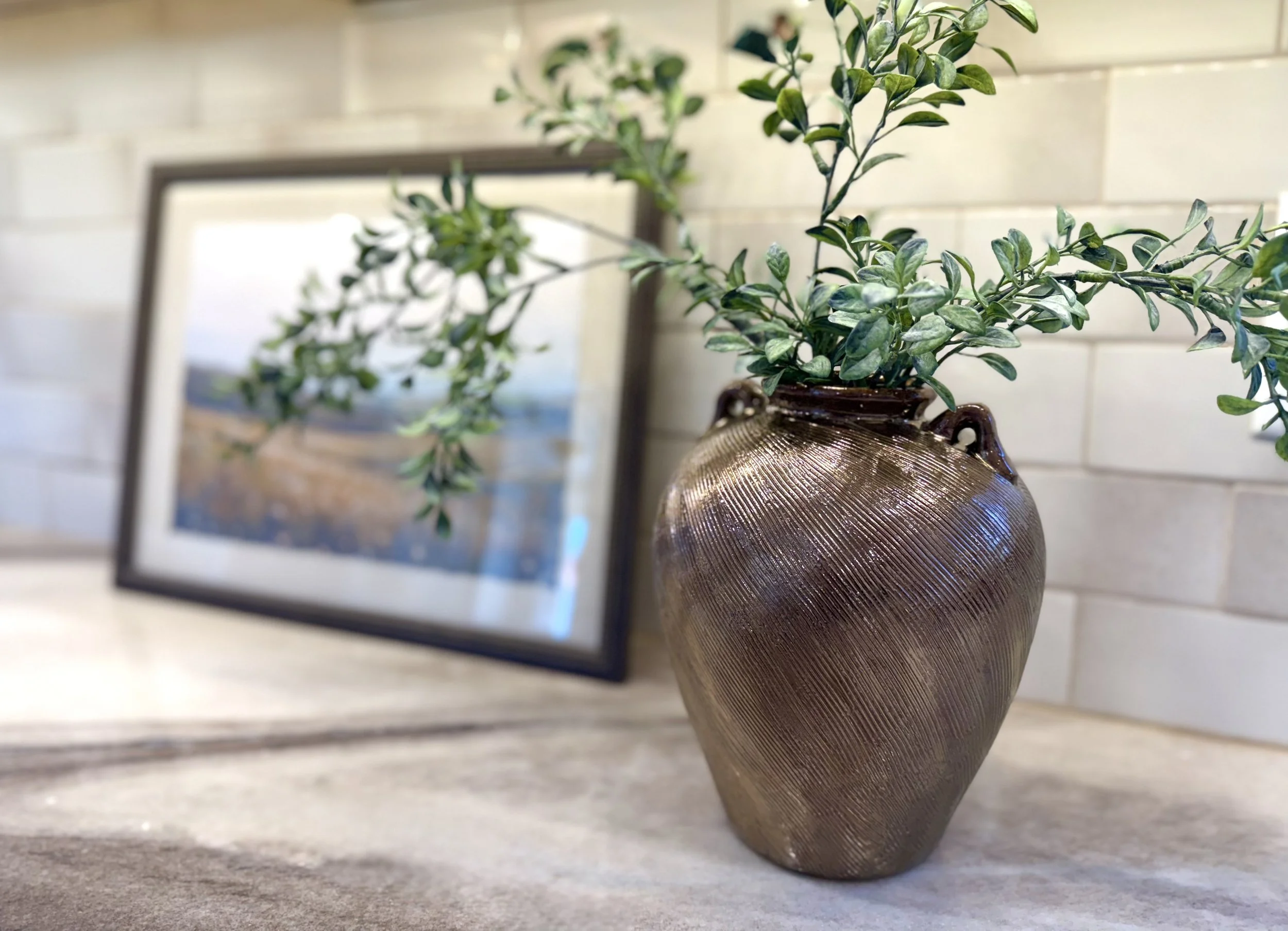 Decorative bronze colored ceramic vase styled with soft greenery on a quartz brown and cream colored countertop, layered with neutral textures and brown framed artwork in the background.