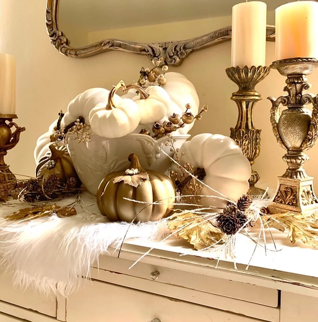 Decorative pumpkins and fall-themed items on a white dresser, including white and gold pumpkins, dried leaves, pinecones, candles, and ornamental branches, with an ornate mirror and candleholders in the background.