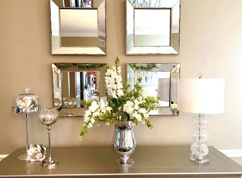 A gray sideboard with a silver vase of white flowers, a clear lamp, and decorative silver and glass bowls, with four square mirrors hanging above on a beige wall.