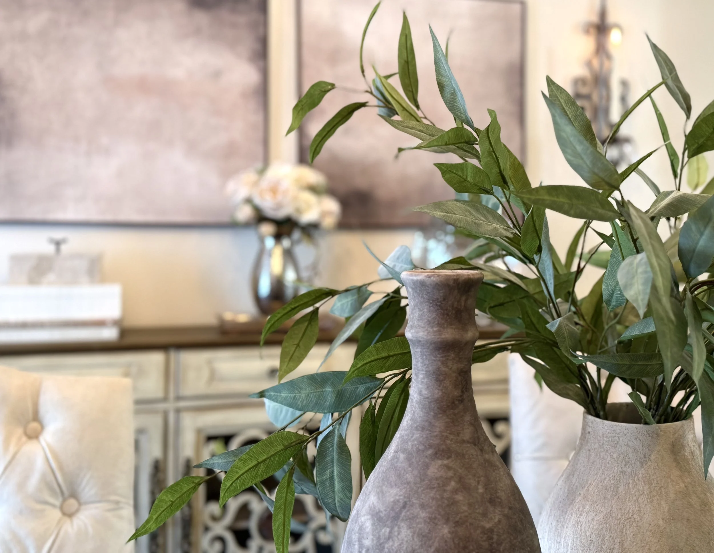 A close-up view of a professionally styled dining room featuring neutral ceramic vases filled with lush greenery on a wooden dining room table. The space showcases soft, layered textures, warm neutral tones, and elegant abstract, modern wall art plan