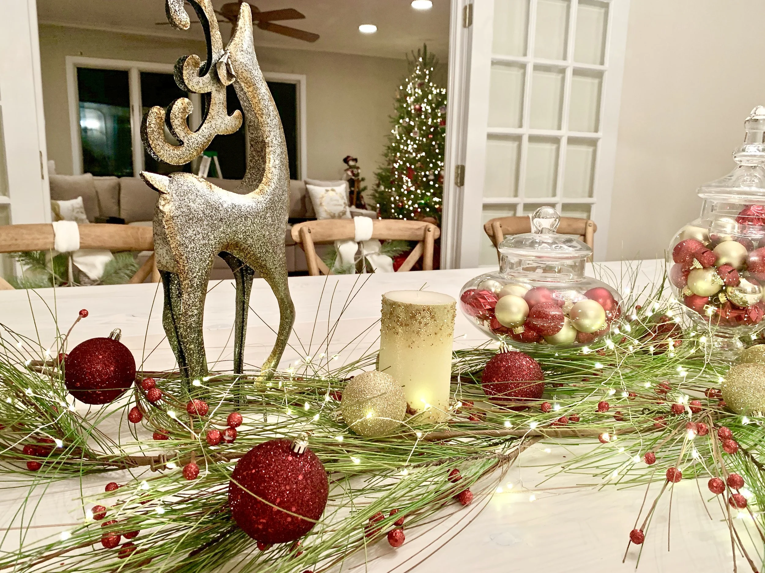 Decorated Christmas table with a glittery reindeer figurine, red and gold ornaments, pine branches with red berries, and a yellow candle, with a Christmas tree and living room in the background.