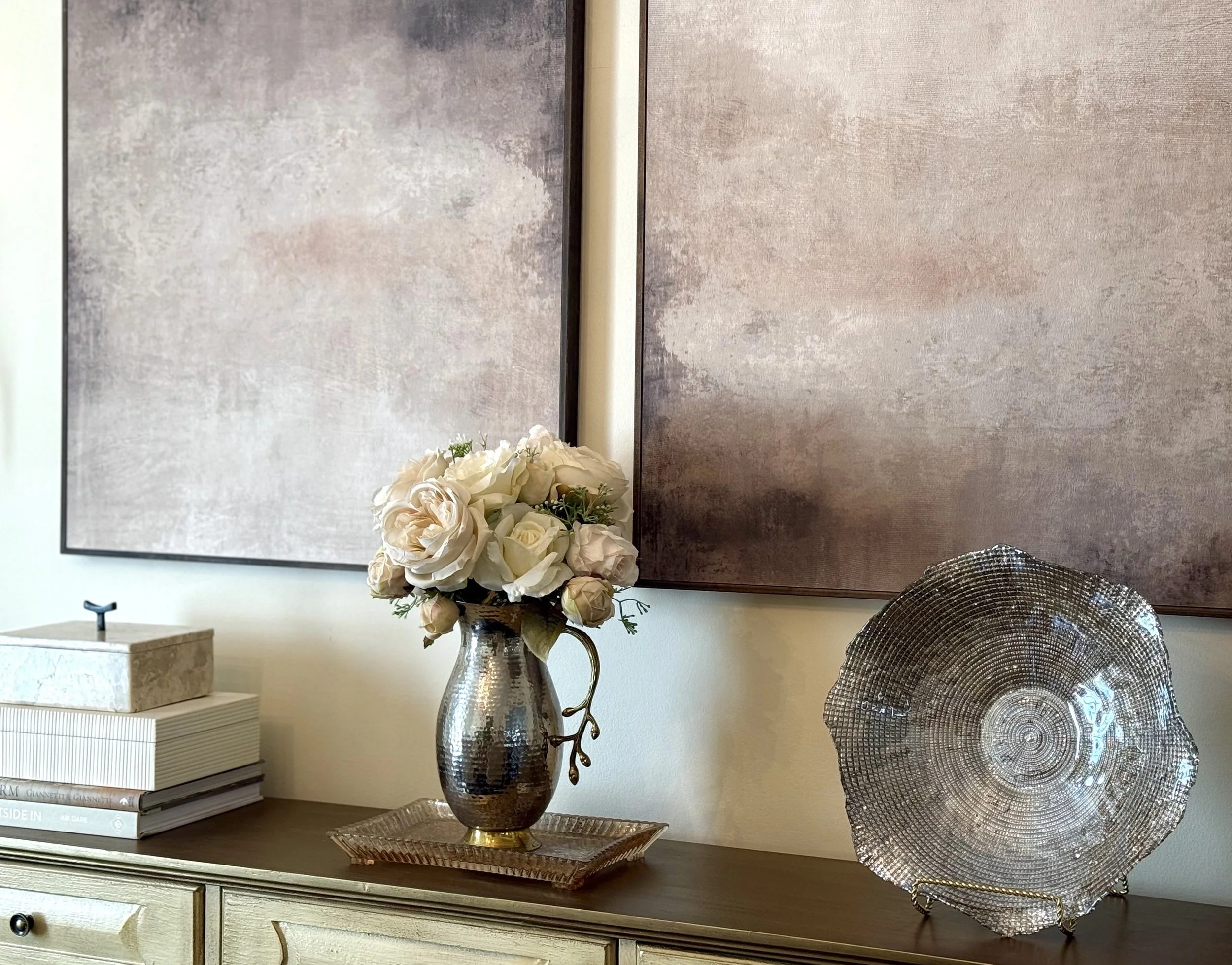 A close-up view of a professionally styled dining room console featuring soft, layered textures, warm neutral tones, and elegant abstract, modern wall art. The distressed wooden console displays a marble decorative box siting on decorative books, a p