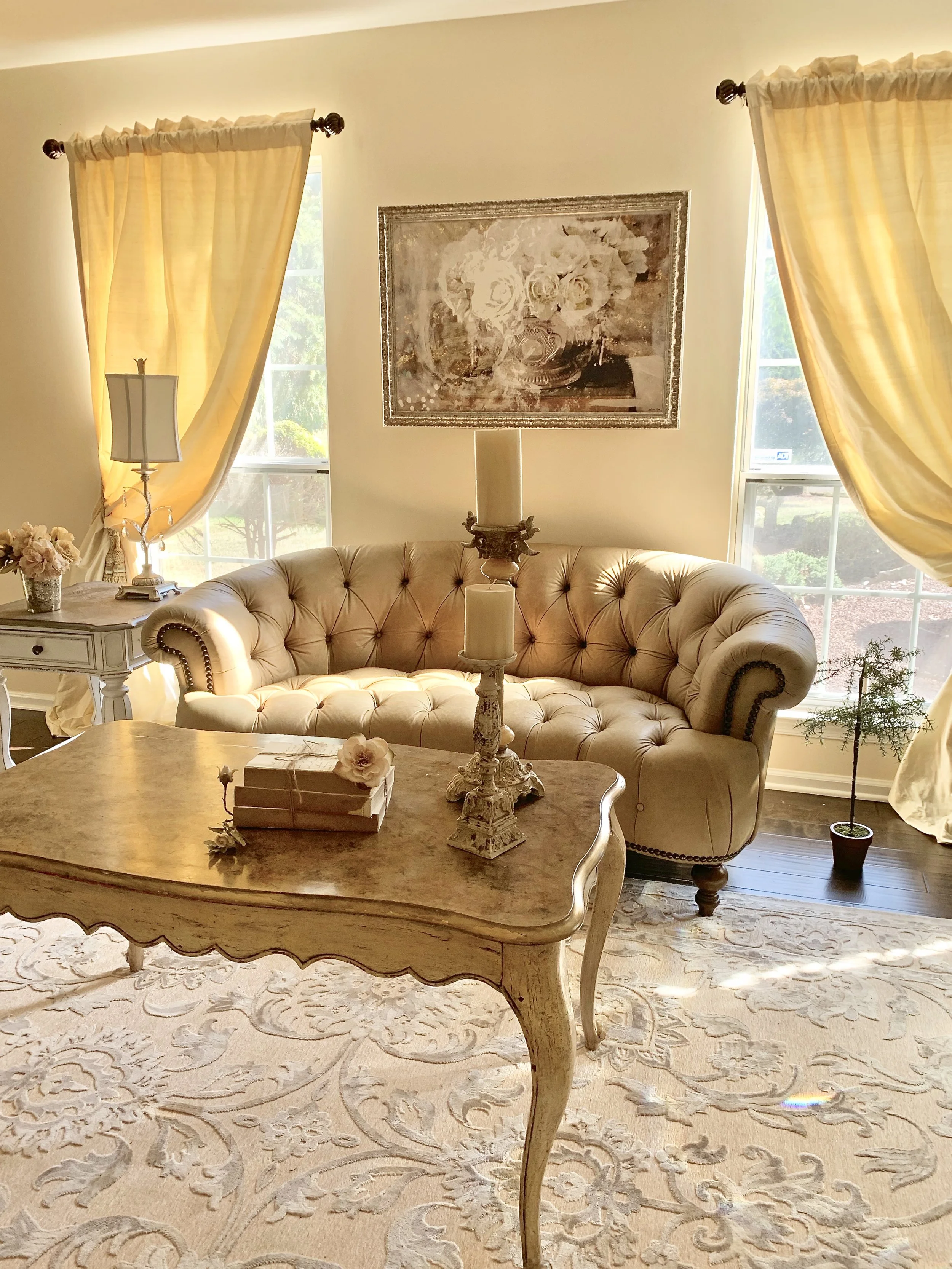 Living room with beige tufted sofa, cream curtains, floral artwork, table with candles and a small potted tree, bright daylight