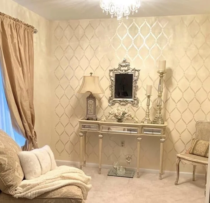 Elegant living room with a chandelier, patterned wallpaper, a cream-colored truck, table with decorative items, mirror, and lamps.