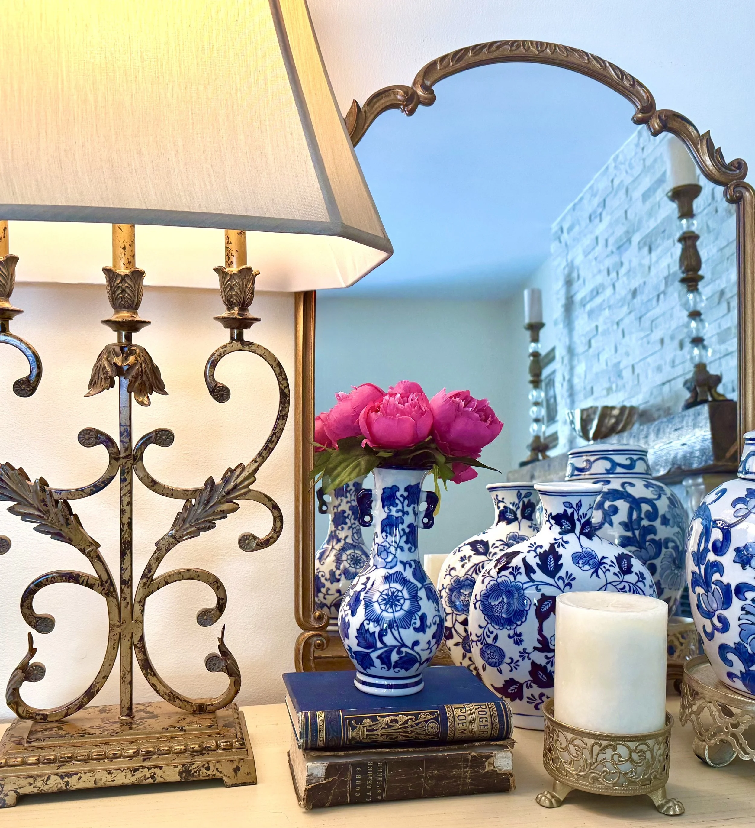 Professionally curated decorative vignette showcasing blue and white floral pottery, fuchsia flowers, stacked blue books, large gold arched mirror, gold pillar candlestick, tall gold candle, white shag accent on a wooden rustic mantle with beige and 