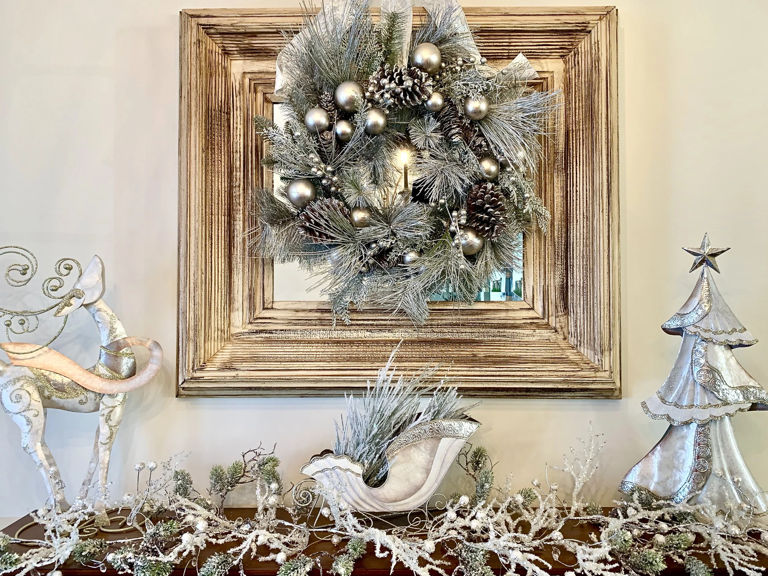 Decorative Christmas display featuring a wreath with silver ornaments and pinecones hanging inside a wooden frame on the wall. On a table below, there is a white reindeer figurine with gold accents, a white sleigh with ornaments, a white Christmas tr