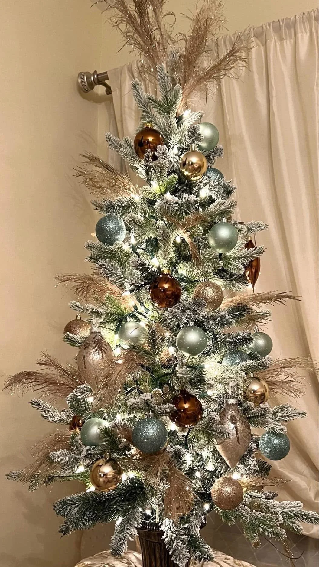 Decorated Christmas tree with white snow-like flocking, gold, silver, and teal ornaments, and feathers in a room with beige walls and white curtains.