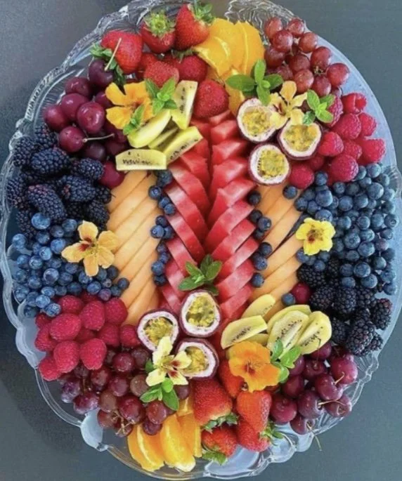 A large round platter filled with an assortment of sliced and whole fresh fruits arranged in a colorful pattern. Fruits include strawberries, grapes, blueberries, blackberries, raspberries, passion fruits, passion fruit halves, orange slices, melon s
