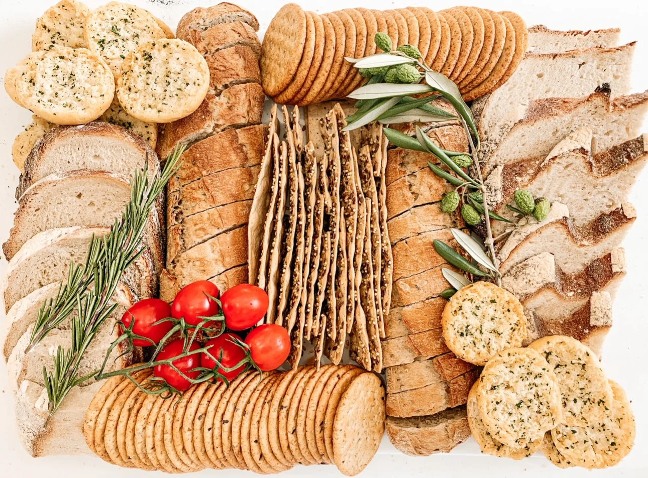 An assortment of bread, crackers, tomatoes, and herbs on a white surface, including sliced bread, rice crackers, tomato clusters, and sprigs of rosemary and green herbs.
