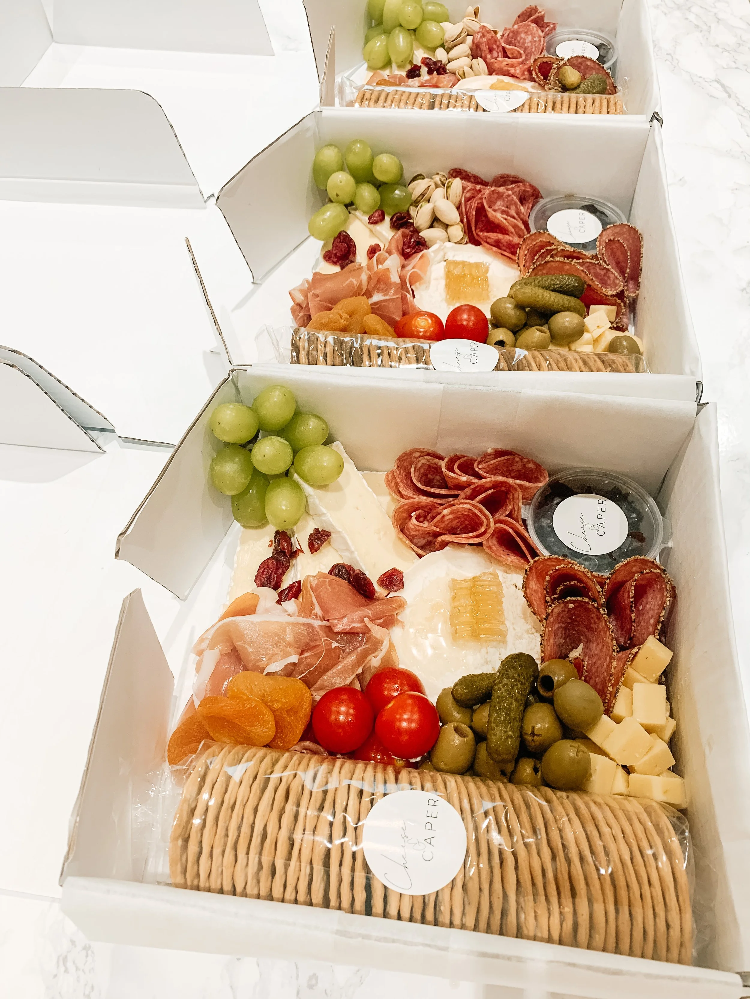 Three white boxes of assorted charcuterie, cheese, grapes, cherry tomatoes, crackers, and pickles.