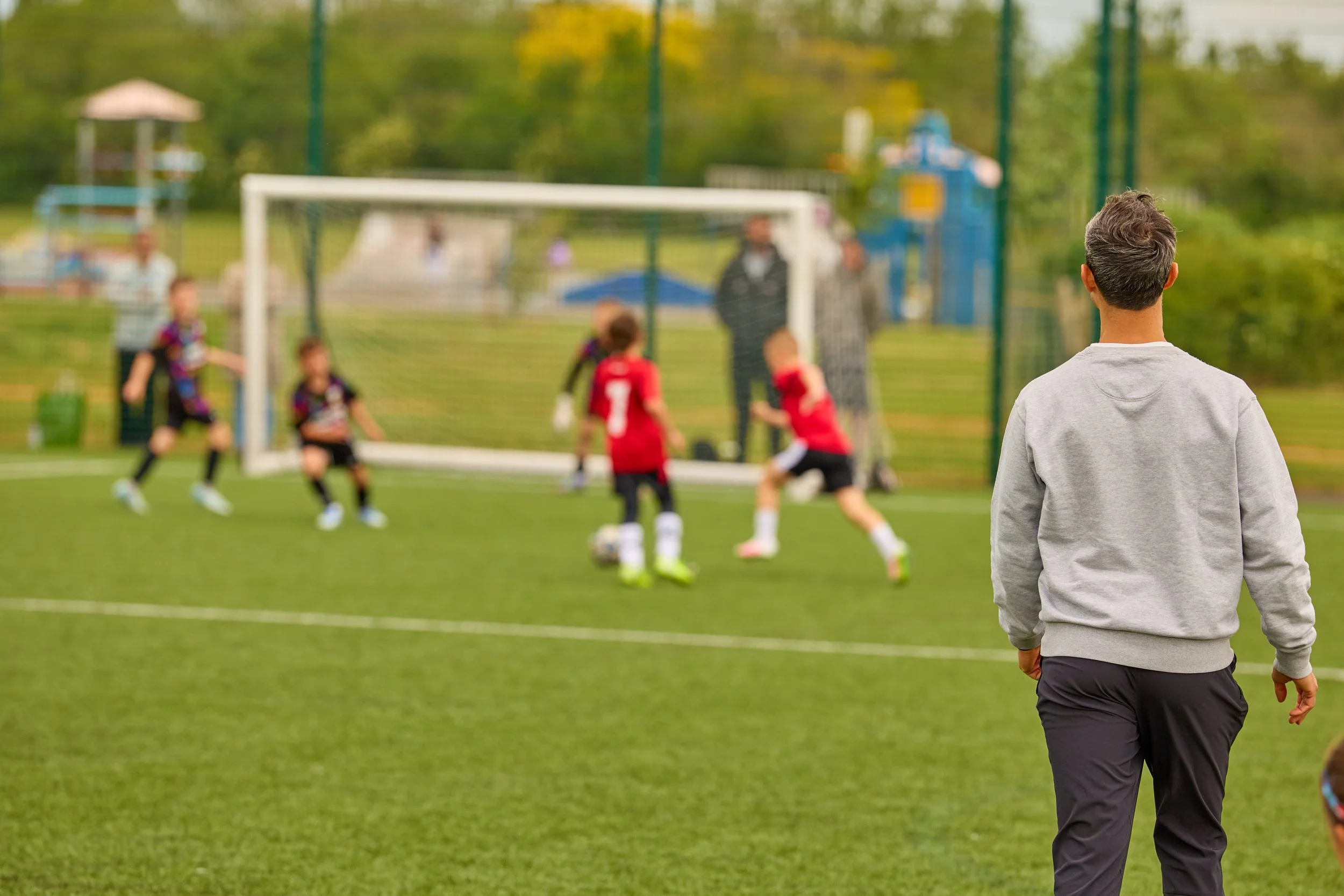 A man in a gray hoodie watching a children’s soccer game on a green field with a playground and trees in the background.
