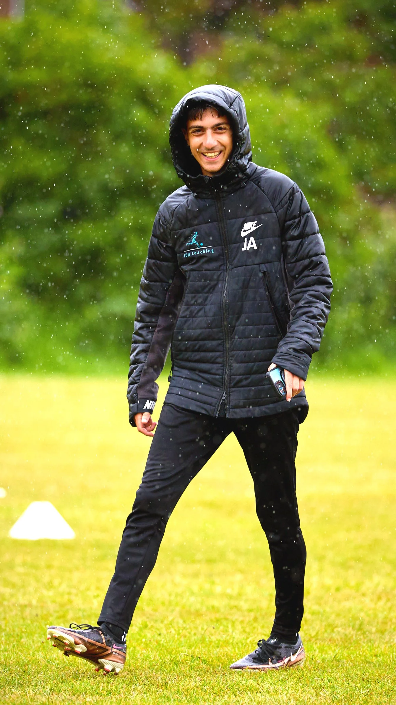 A young man in sportswear smiling outdoors in the rain on a grassy field.