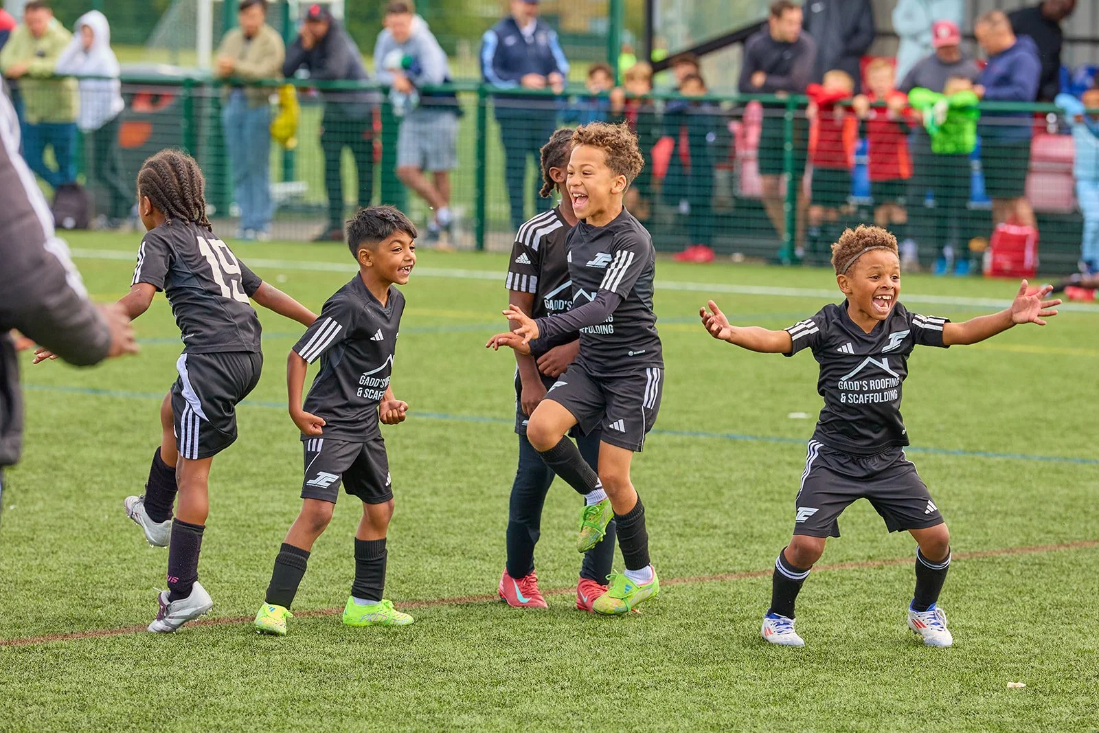 Young children in black soccer uniforms celebrating on a soccer field with spectators watching in the background.