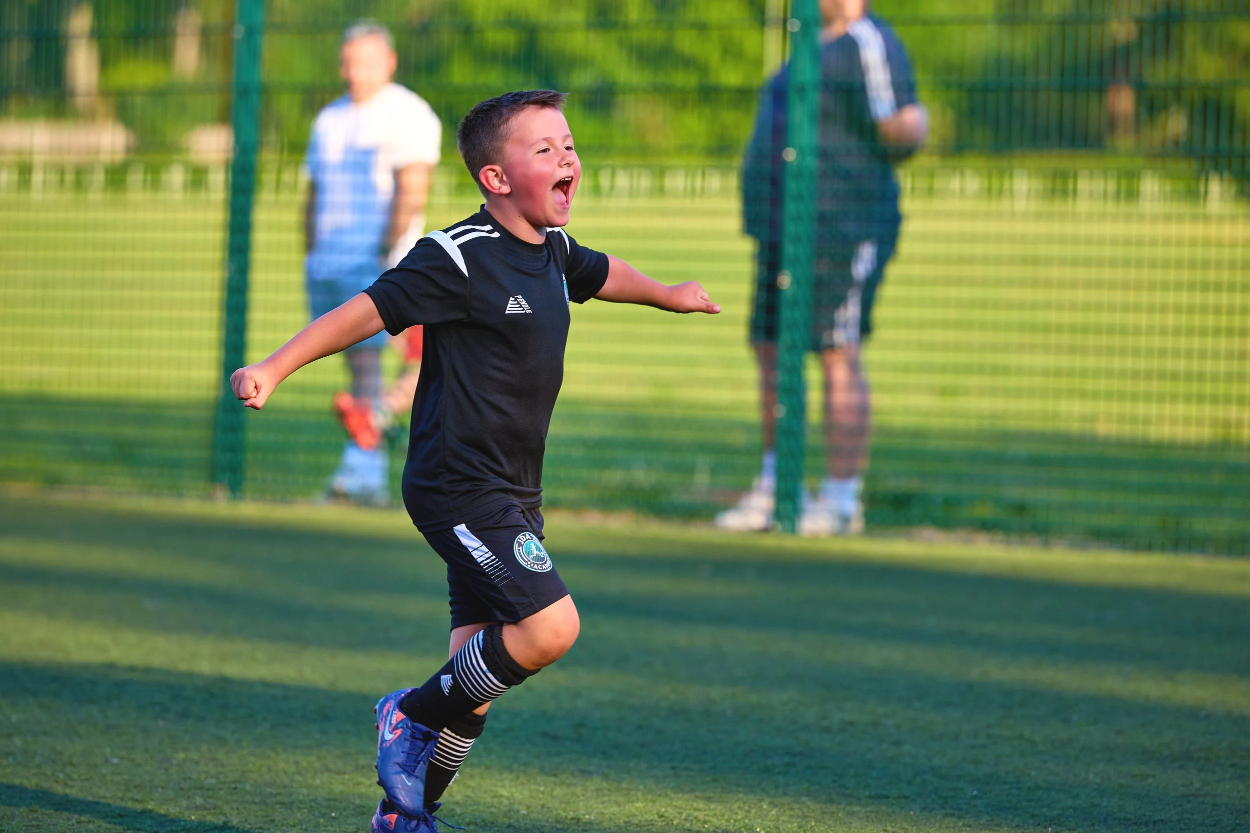 A young boy in a black soccer uniform celebrating a goal on a soccer field, with three adults in the background watching.
