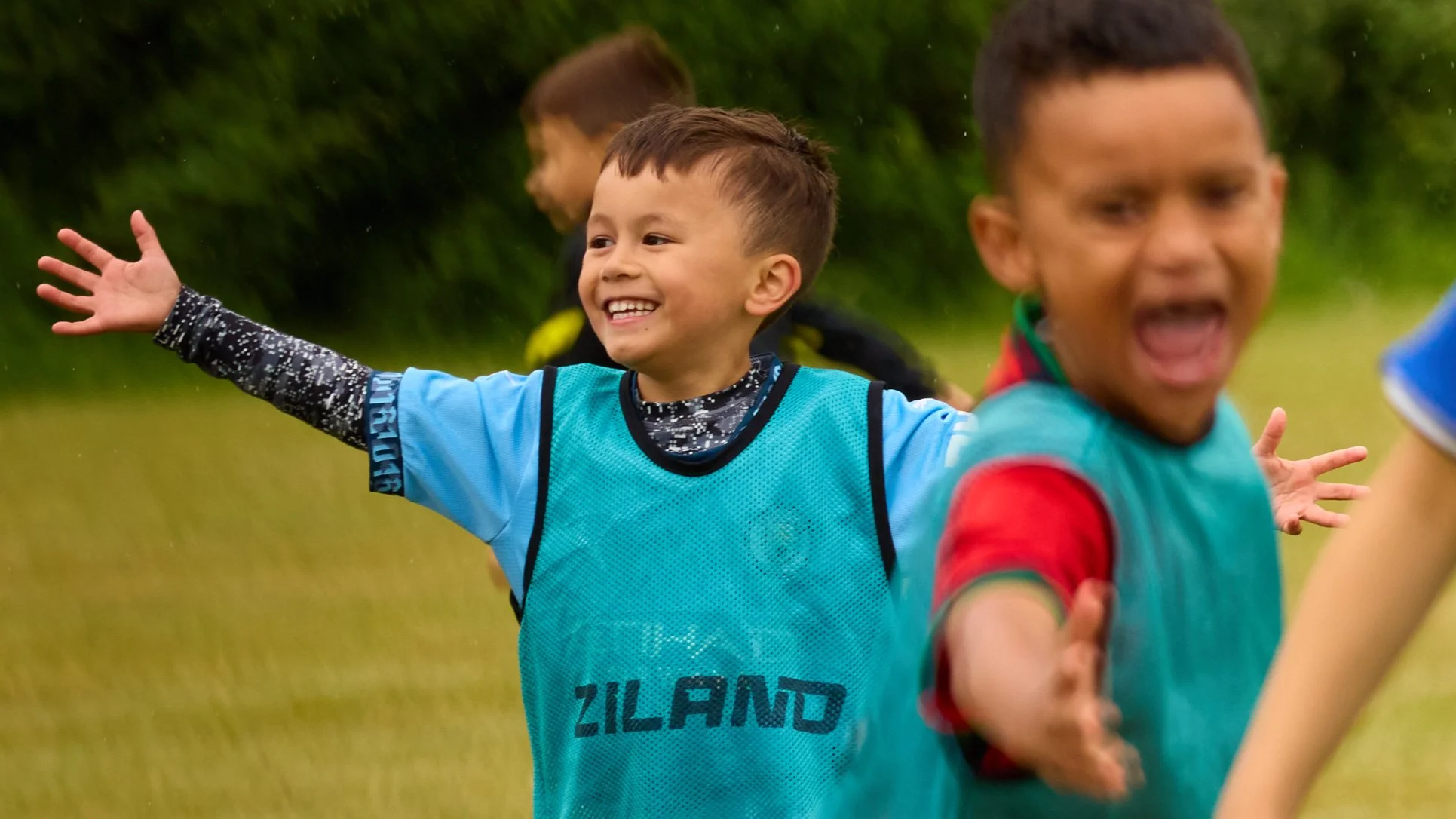 Young boys playing soccer outdoors, smiling, with arms outstretched during a rain shower.