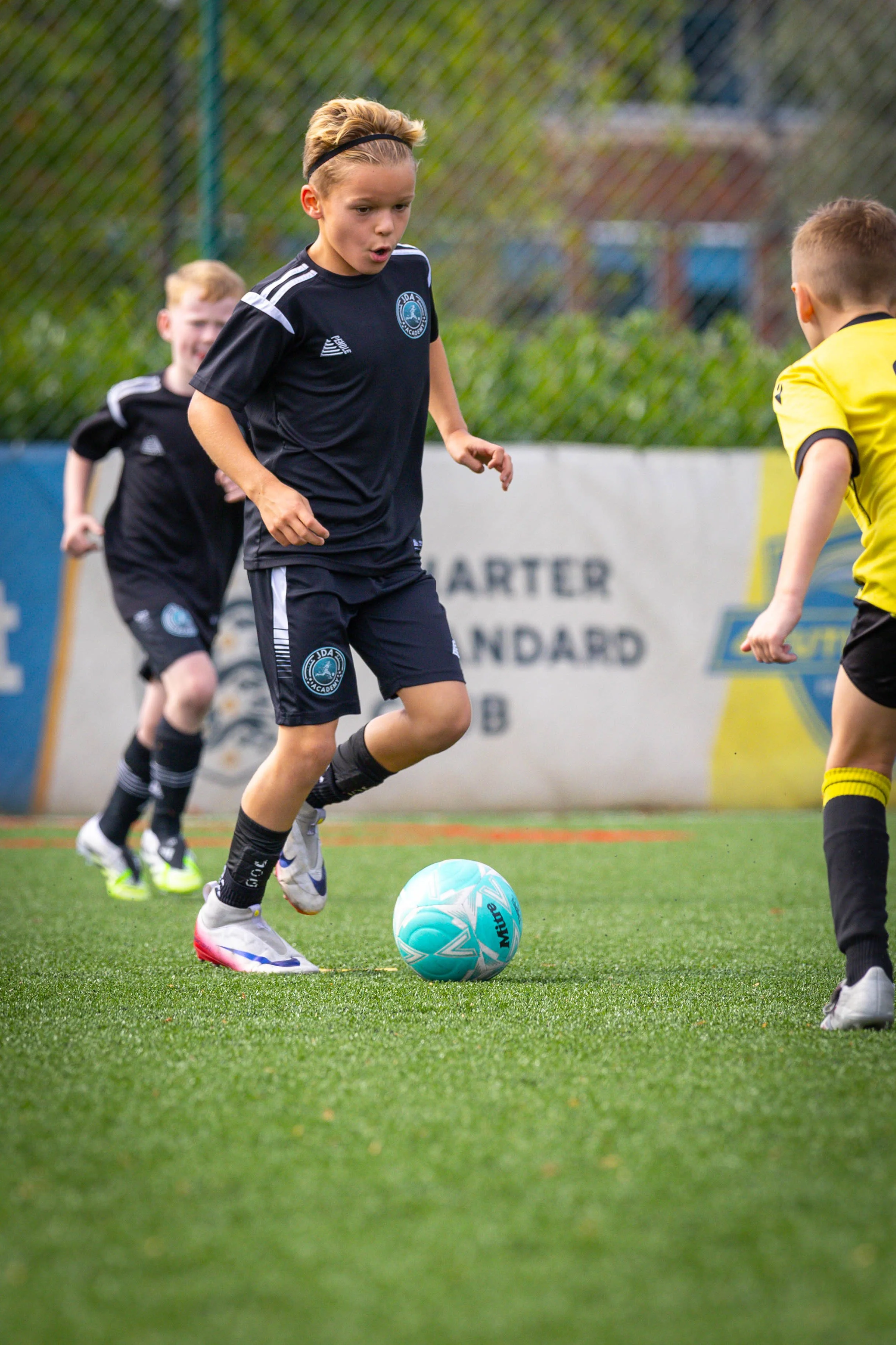 Young boys playing soccer on a field, one in black sports uniform dribbling a blue ball, while others chase behind.