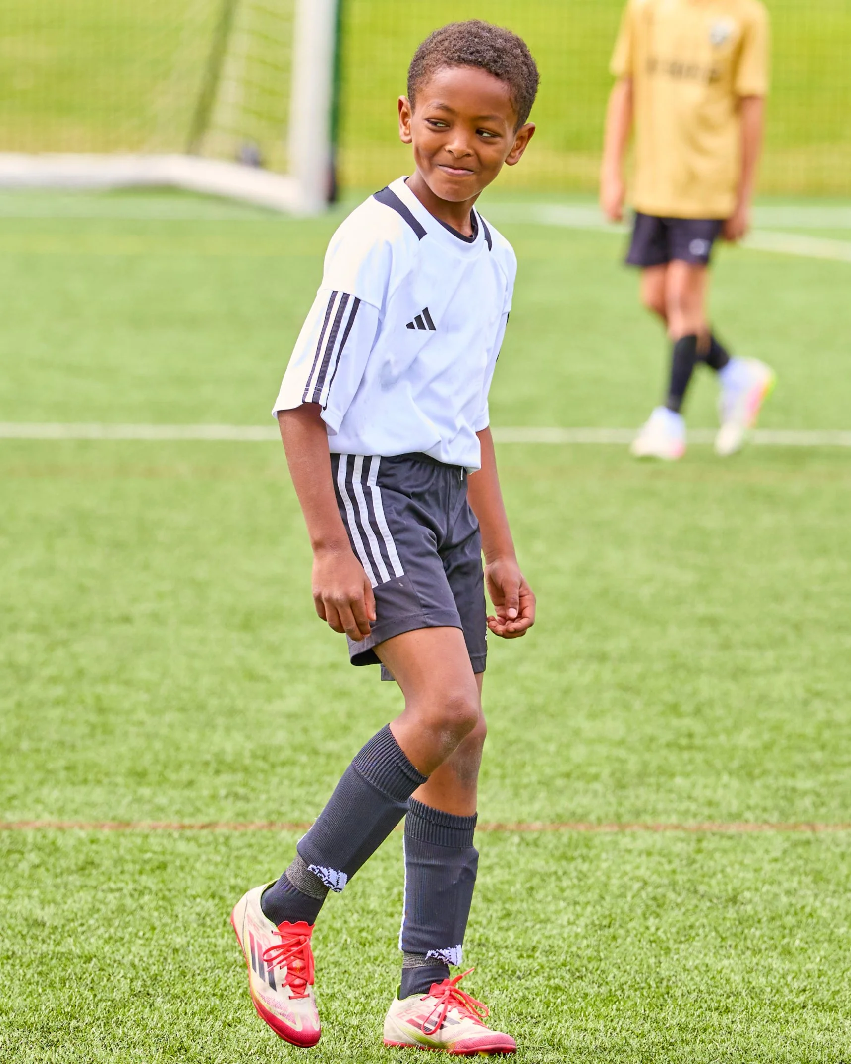 A young boy wearing a white sports jersey, dark shorts, and pink soccer shoes standing on a green soccer field with a goal in the background. Another child is blurred in the background.