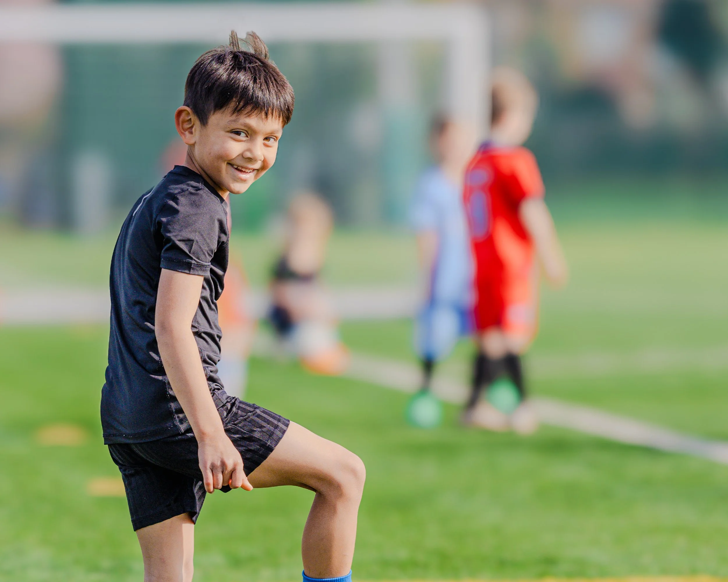A young boy in a black sports uniform, smiling and standing on a soccer field during a game or practice, with other children blurred in the background.