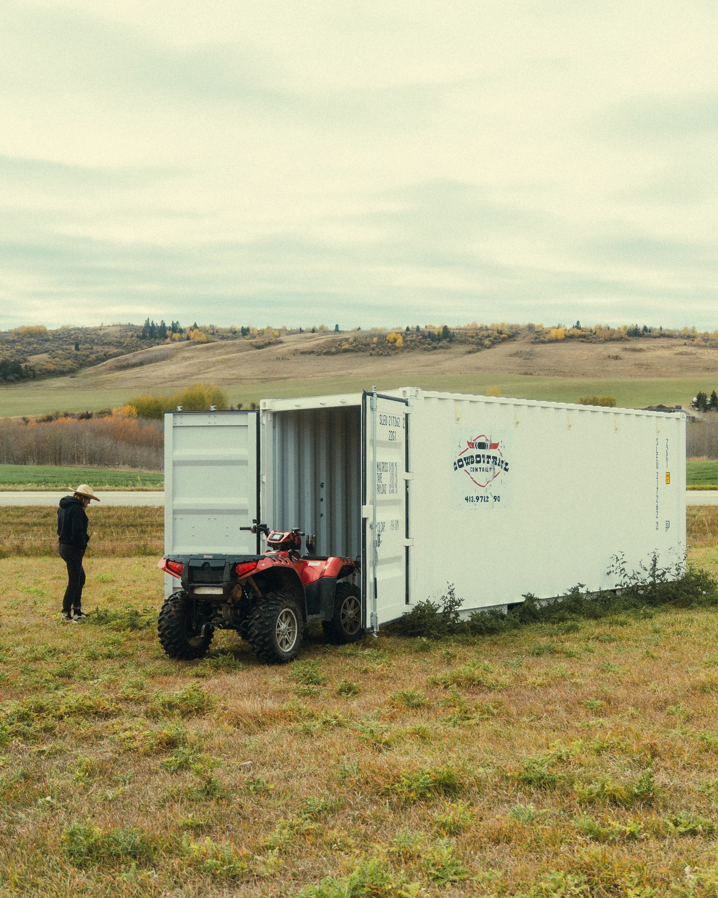 Person standing next to a red ATV inside a large white shipping container on a grassy field with rolling hills in the background.