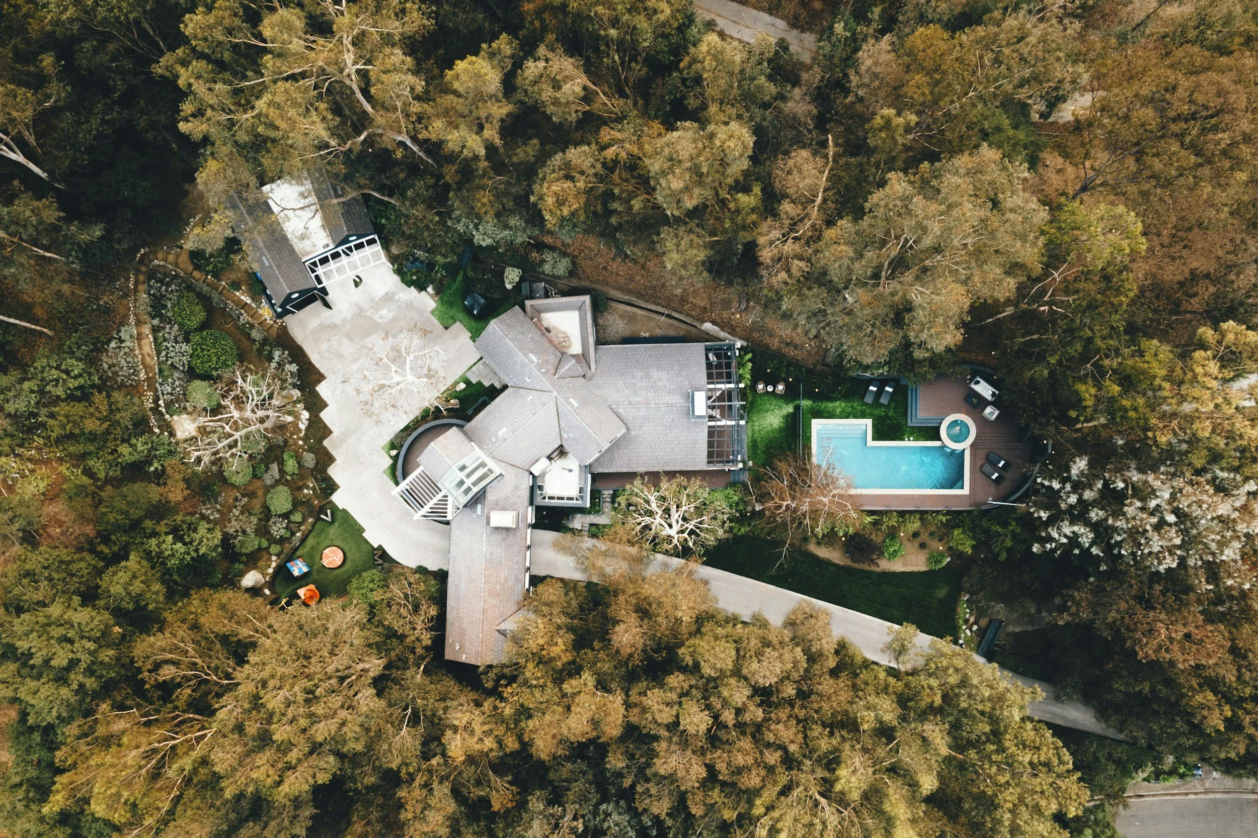 Aerial view of a house surrounded by trees, with a large driveway, a swimming pool, and a landscaped yard.