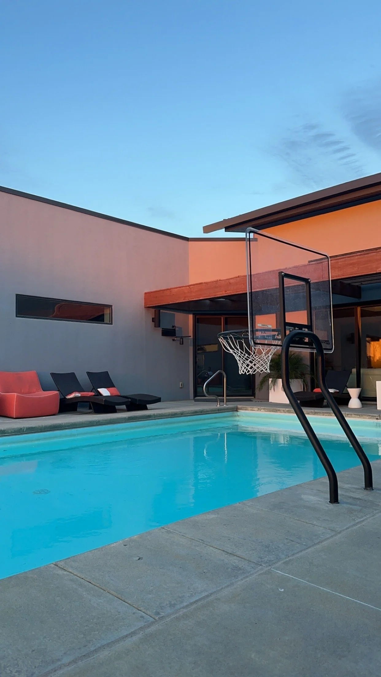 Modern backyard with a swimming pool, lounge chairs, a basketball hoop, and a patio with modern architecture against a blue sky at sunset.