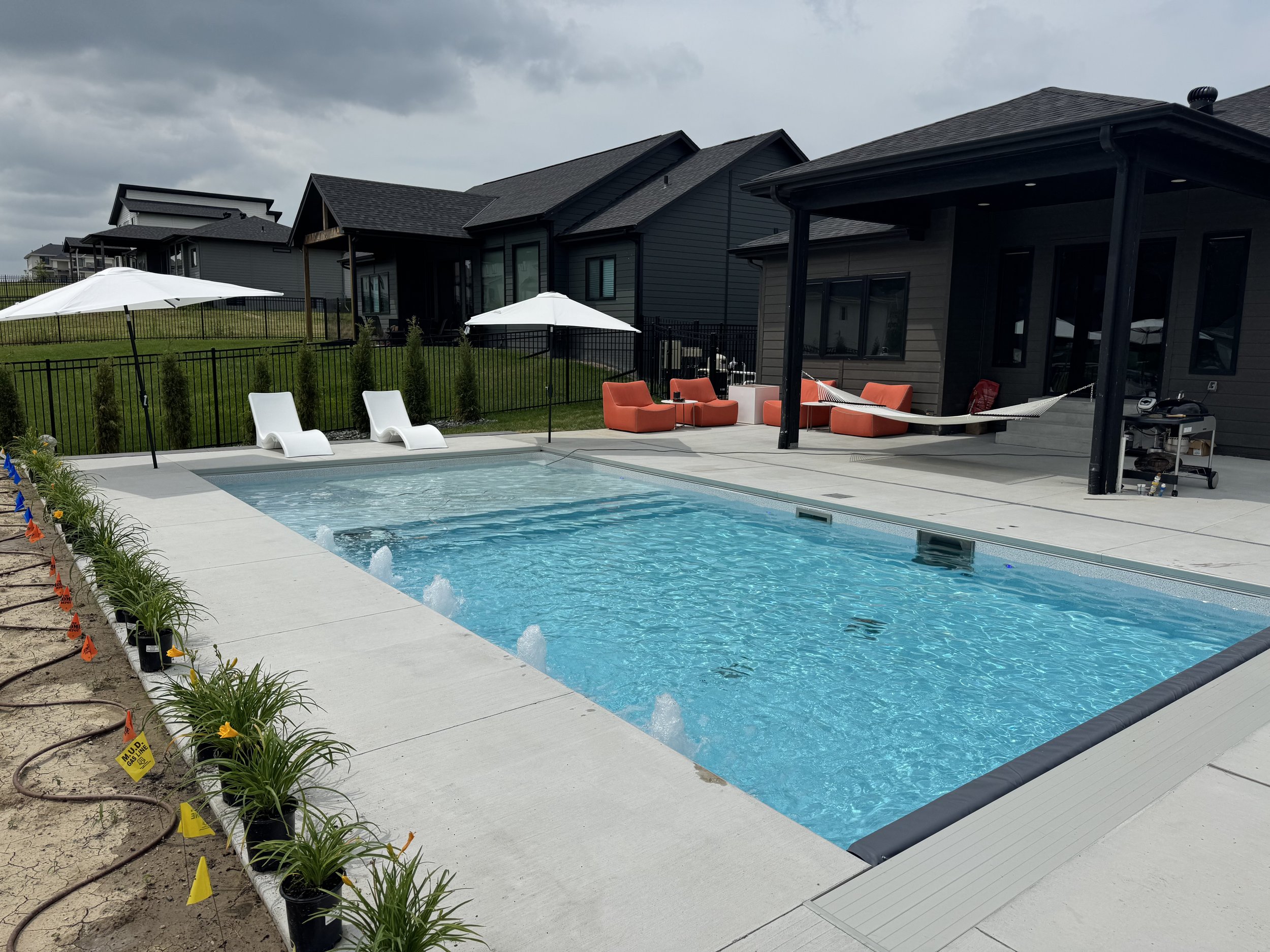 Backyard with a swimming pool, white lounge chairs, umbrellas, outdoor seating, hammock, and a black house with a porch, and neighboring houses in the background.