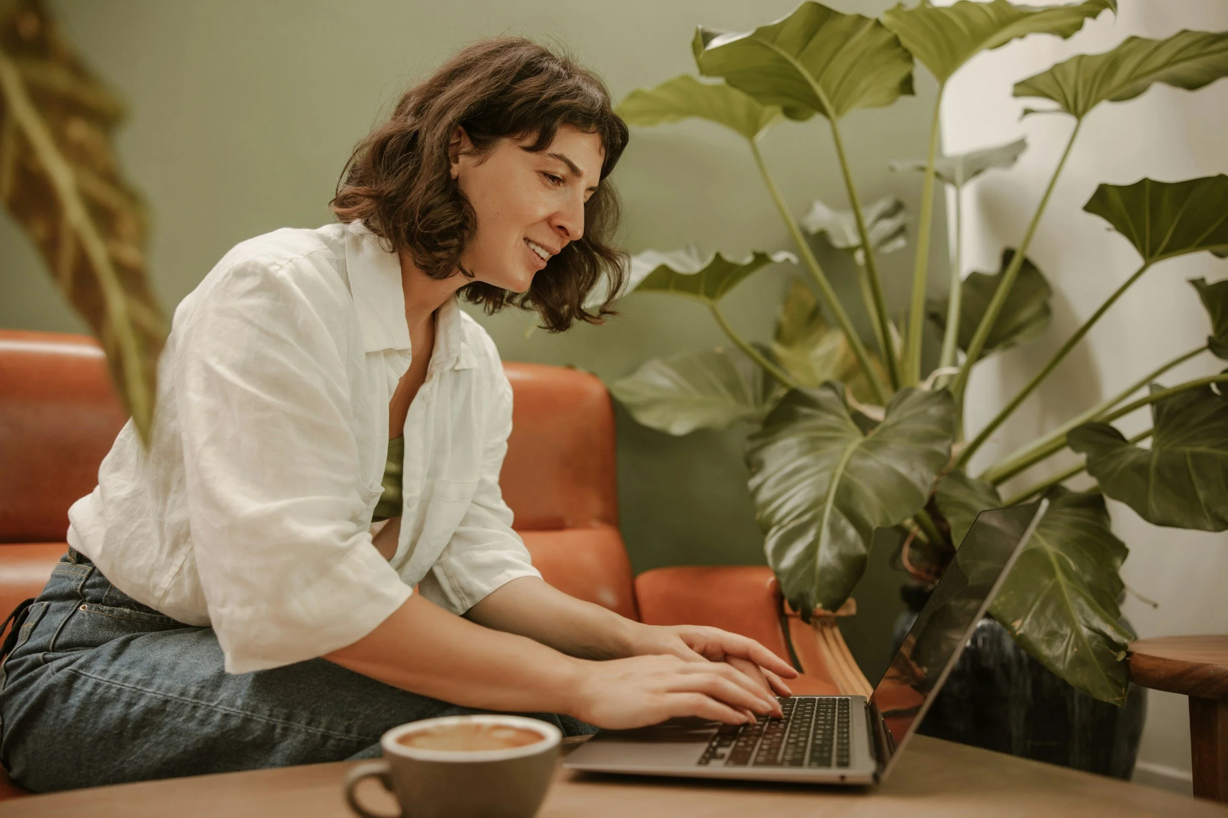 A woman with curly brown hair wearing a white shirt, sitting on an orange couch, smiling and typing on a laptop in a cozy room with green plants.