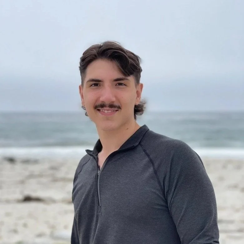 Young man with dark hair and a mustache smiling at the beach, wearing a dark zip-up jacket, with the ocean and cloudy sky in the background.