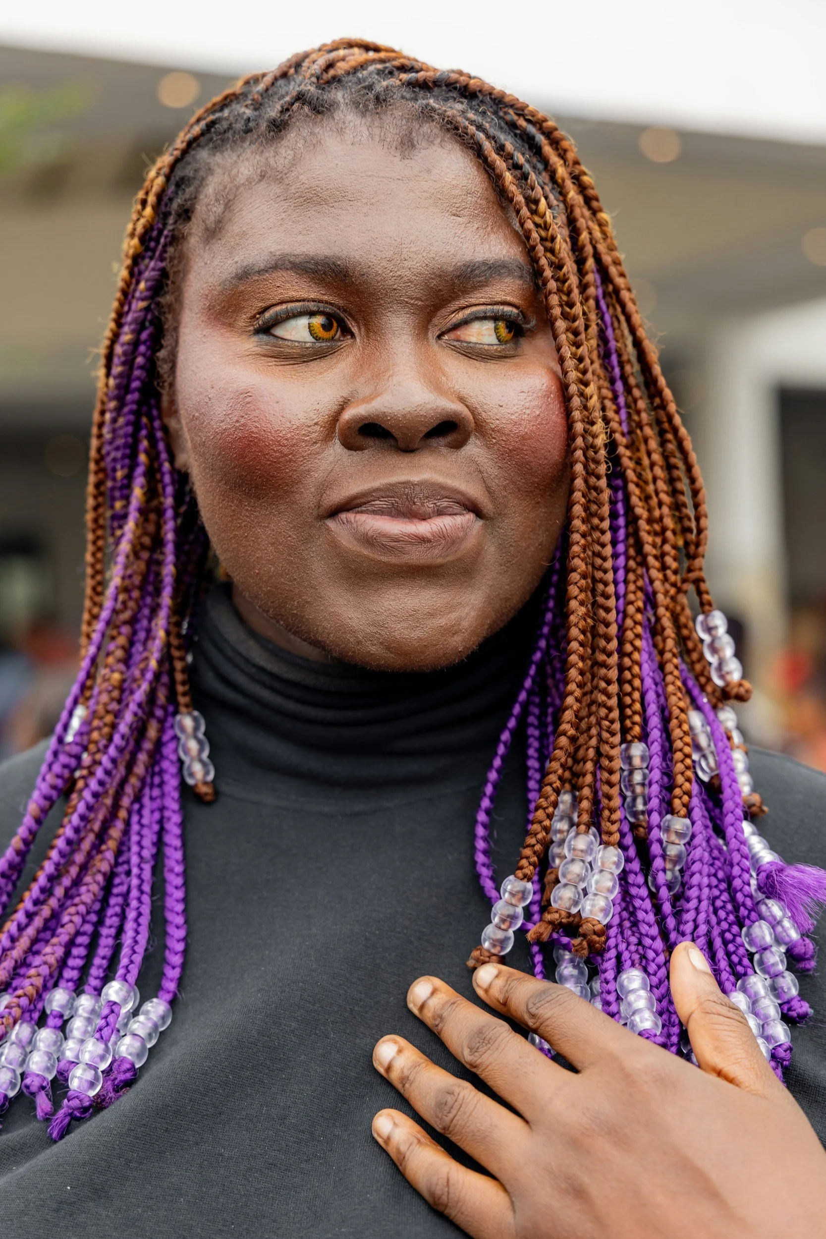 Close-up of a woman with brown skin, yellow eyes, and colorful braided hair, wearing a black top, with her hand on her chest.