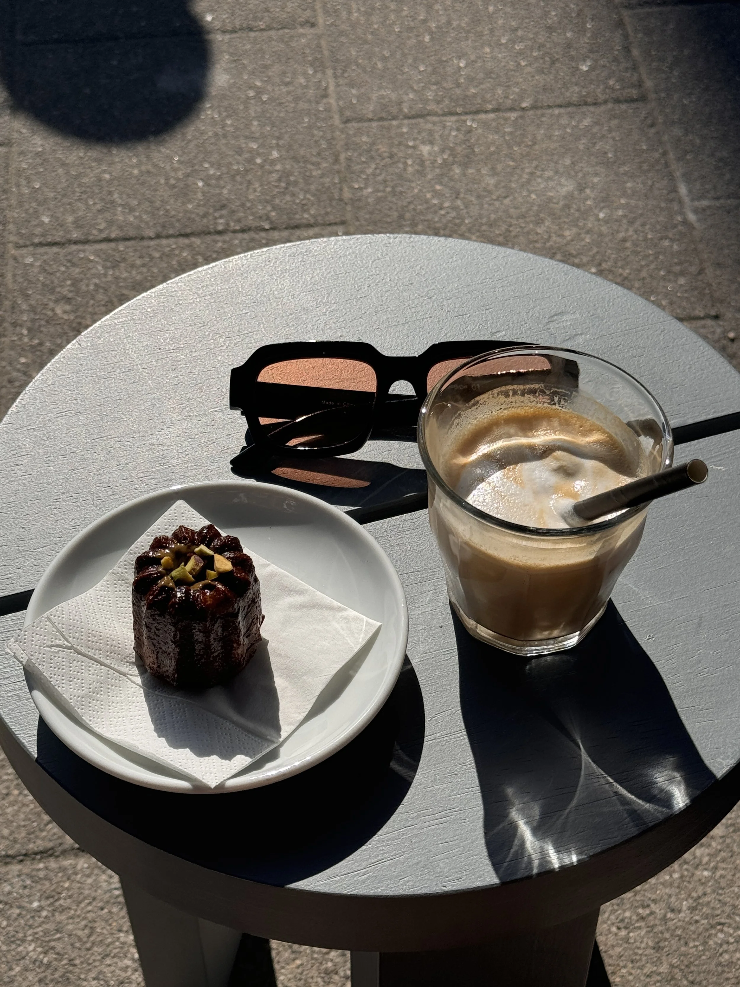 A white table with a slice of cake on a white plate, a glass of iced coffee with a straw, and a pair of black sunglasses placed on the table in sunlight.