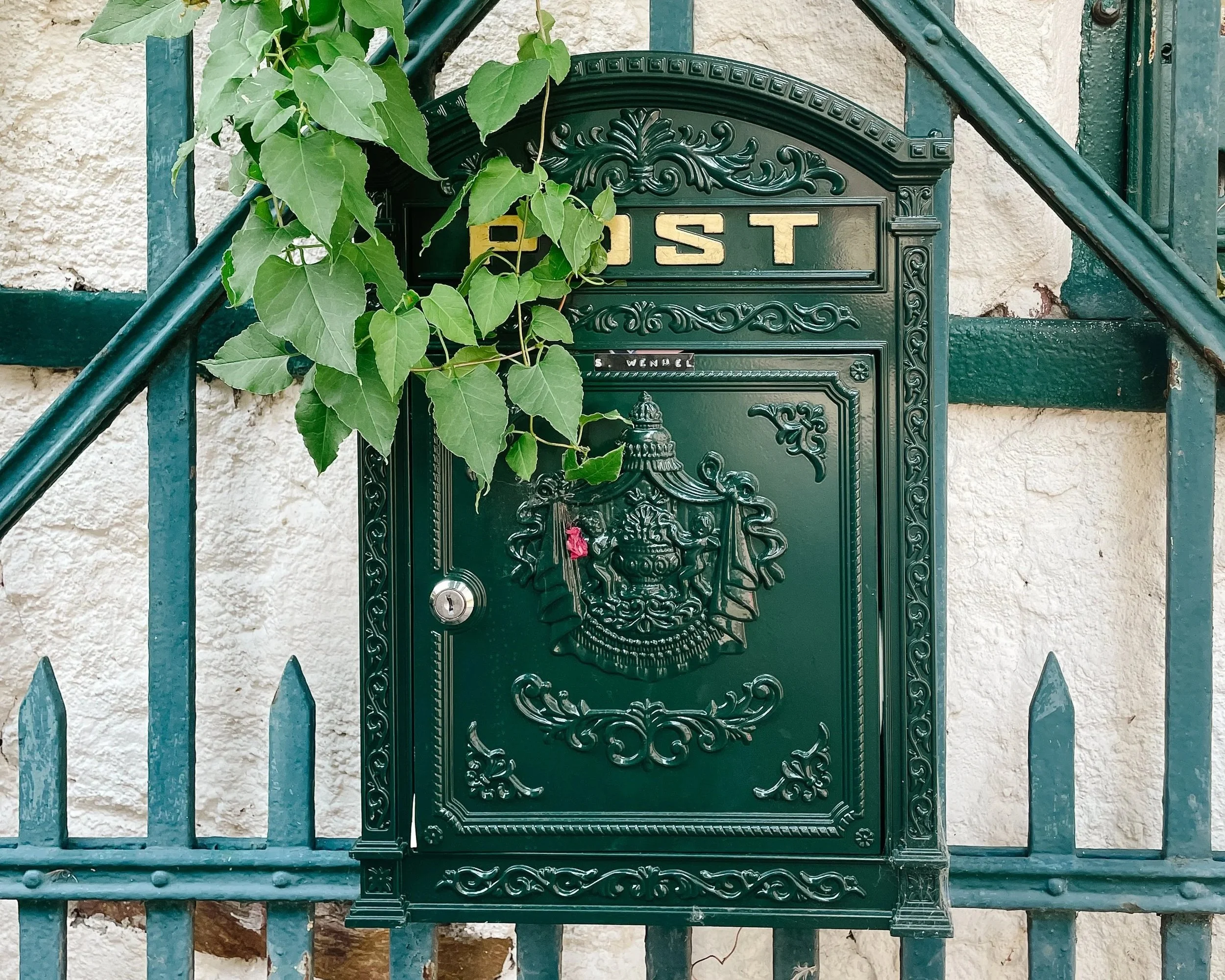 Green decorative mailbox with embossed crest, mounted on a green fence with ivy leaves partially covering it, and a beige textured wall in the background.