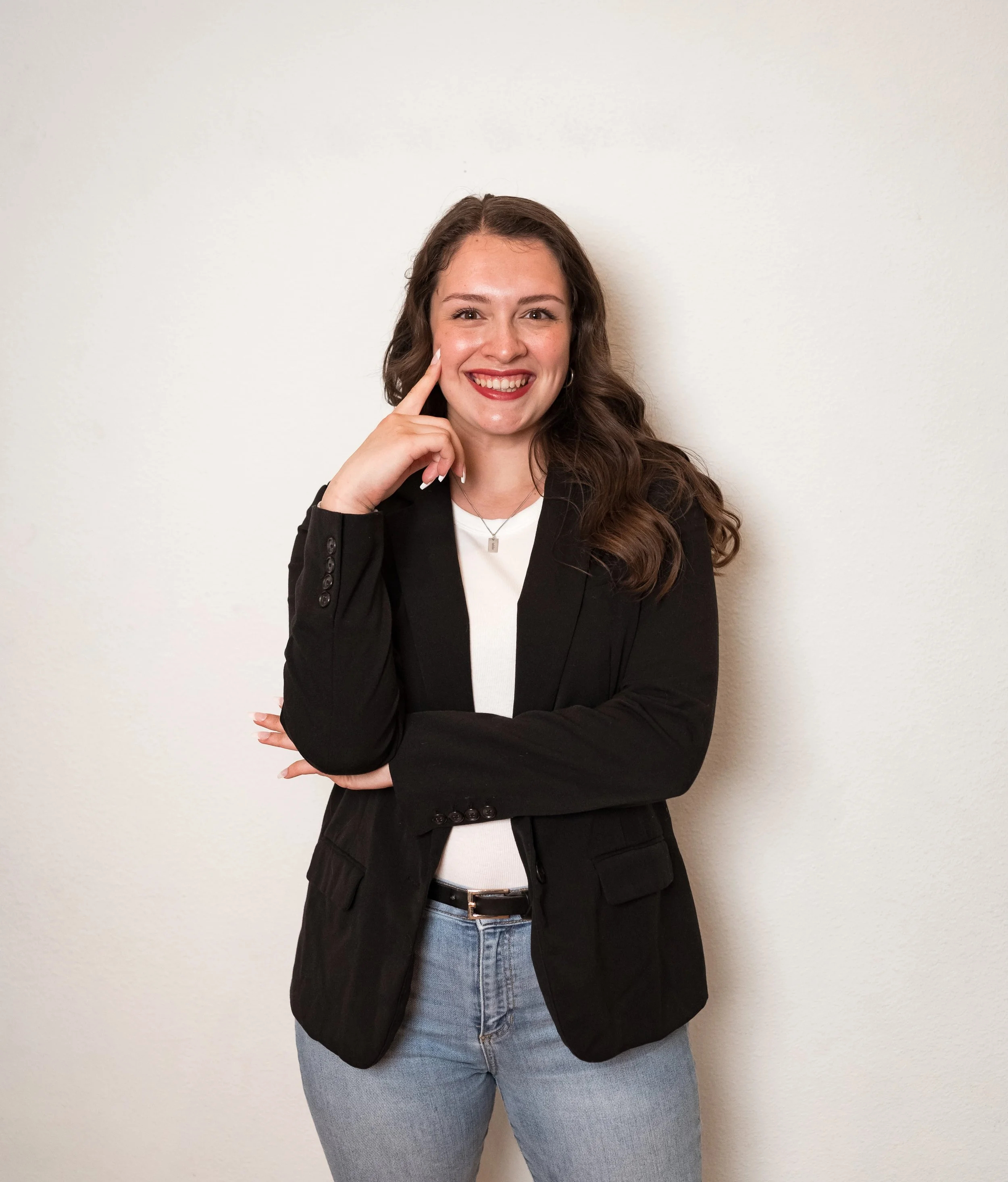 a woman with long wavy brown hair, wearing a black blazer, white top, and light blue jeans, smiling with her hand resting on her cheek against a plain white wall
