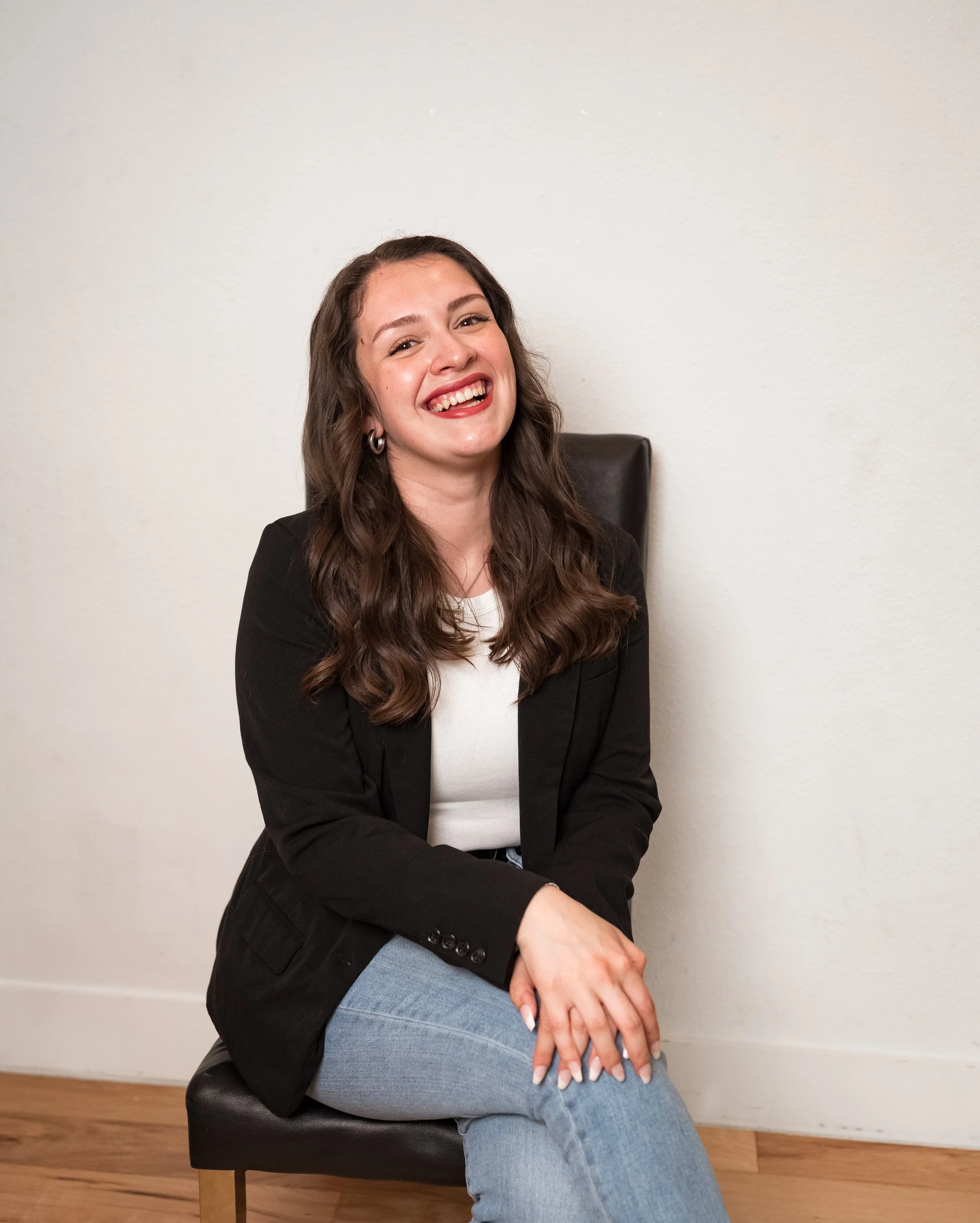 A young woman with long brown hair, wearing a black blazer and light blue jeans, sitting on a black chair against a plain white wall, smiling and looking at the camera.