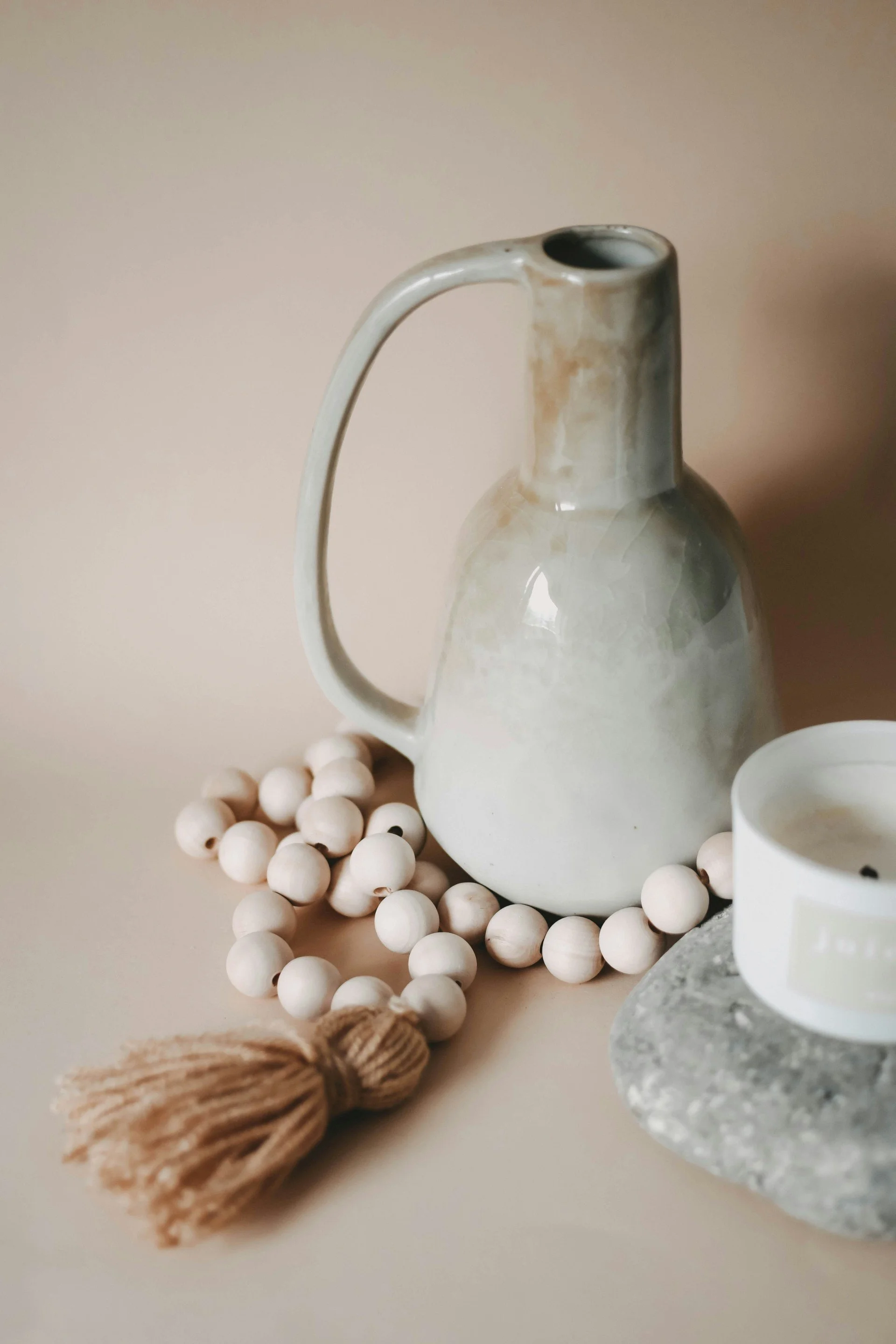 A ceramics jug with a handle and a narrow opening, decorated with earthy glaze tones, surrounded by white wooden beads, a small bundle of beige twine, and a white cup on a smooth surface with a neutral background.