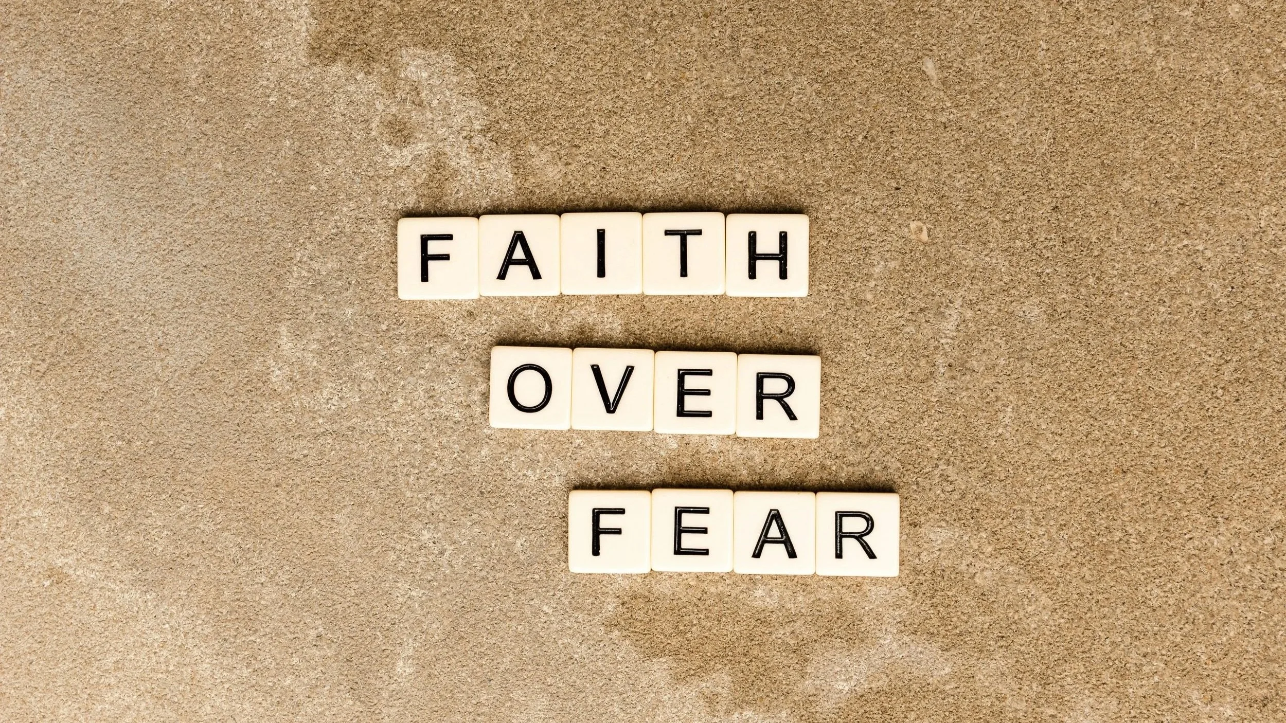 Letters on Scrabble tiles spell out "FAITH OVER FEAR" on a sandy surface.