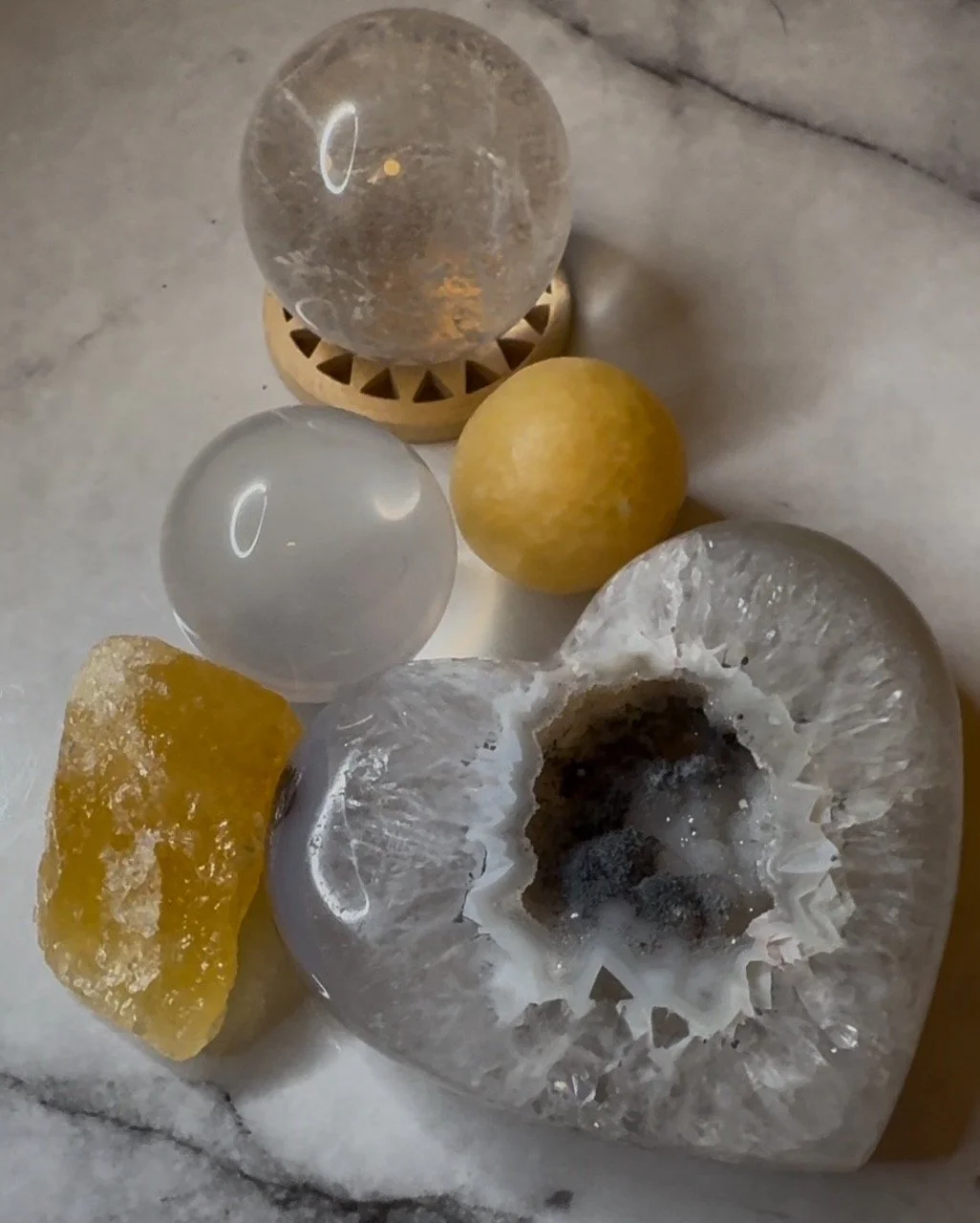 A collection of objects on a marble surface, including a large clear geode with a hollow center, a yellow stone, two polished spherical stones (one clear and one yellow), a yellow crystalline mass, and a small circular wooden stand with a clear glass sphere on top.