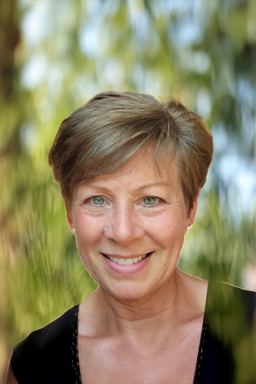 A middle-aged woman with short light brown hair and blue eyes, smiling outdoors with blurry green trees in the background.
