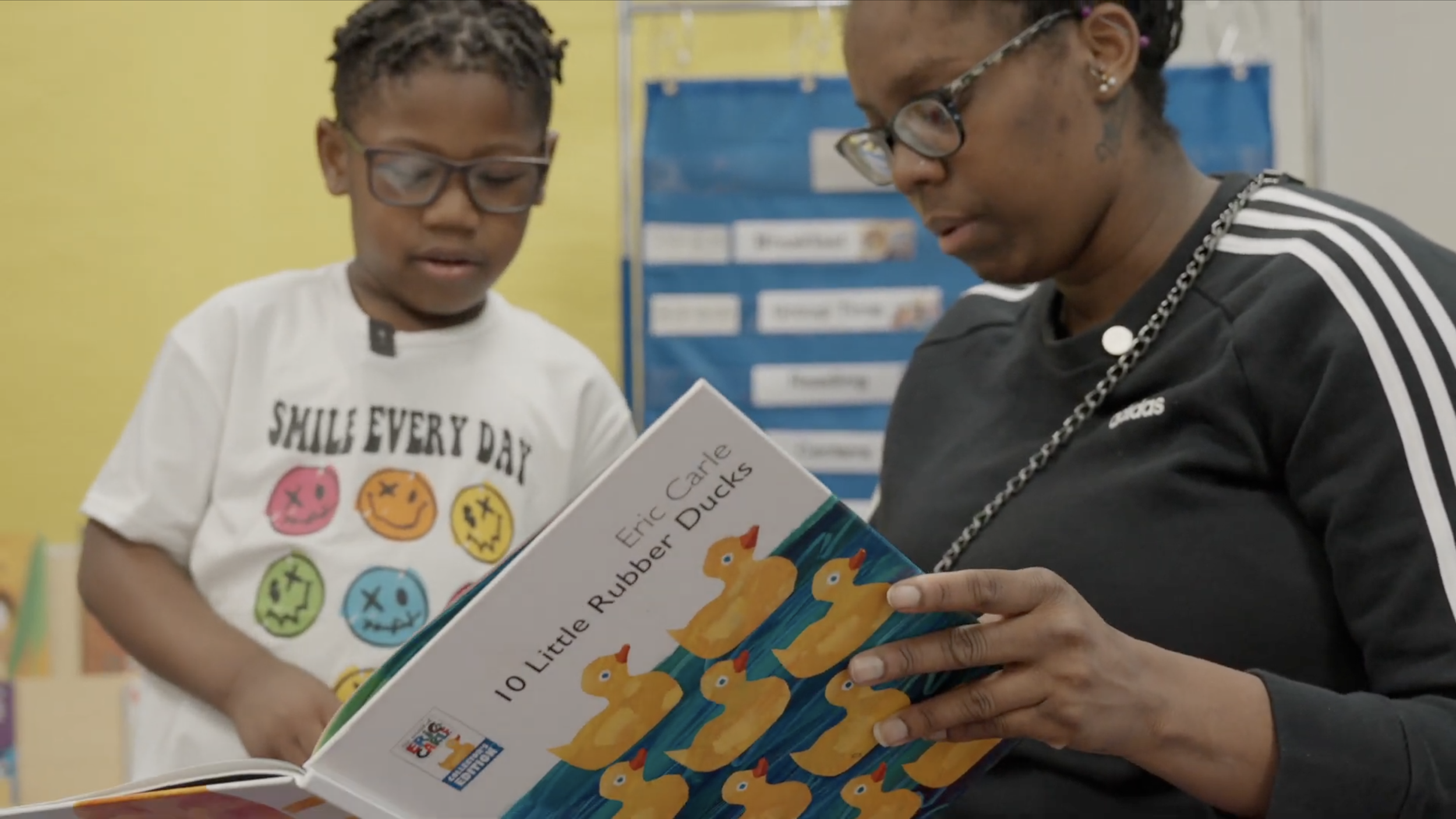 A young boy and a woman reading a colorful children's book titled "10 Little Rubber Ducks" by Eric Carle in a classroom.