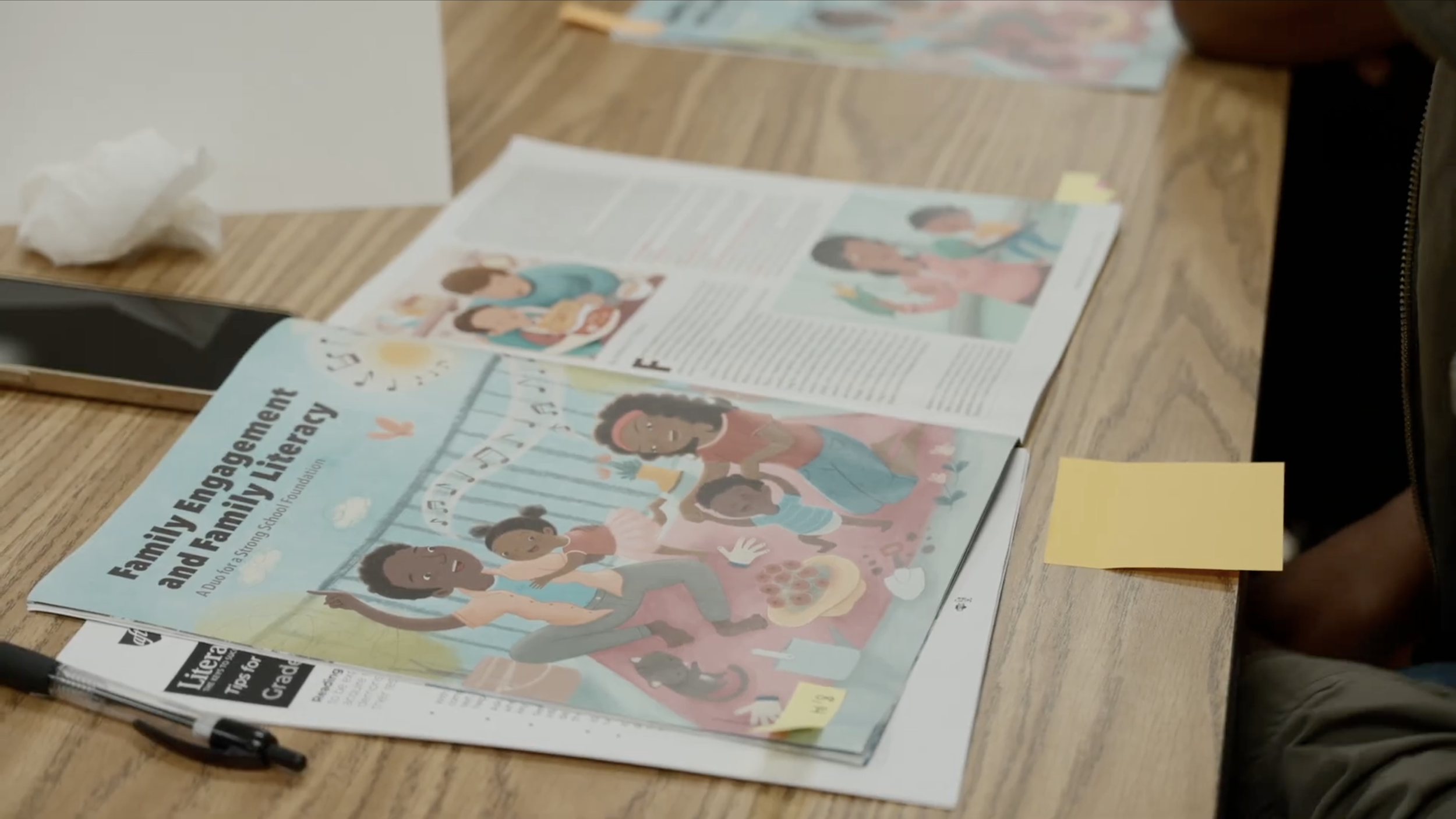 A brochure titled "Family Engagement and Family Literacy showing illustration of a family reading outside with the sun, clouds, and a pink bird, on a wooden table along with a pen, a pencil, a tablet, yellow sticky notes, and papers.