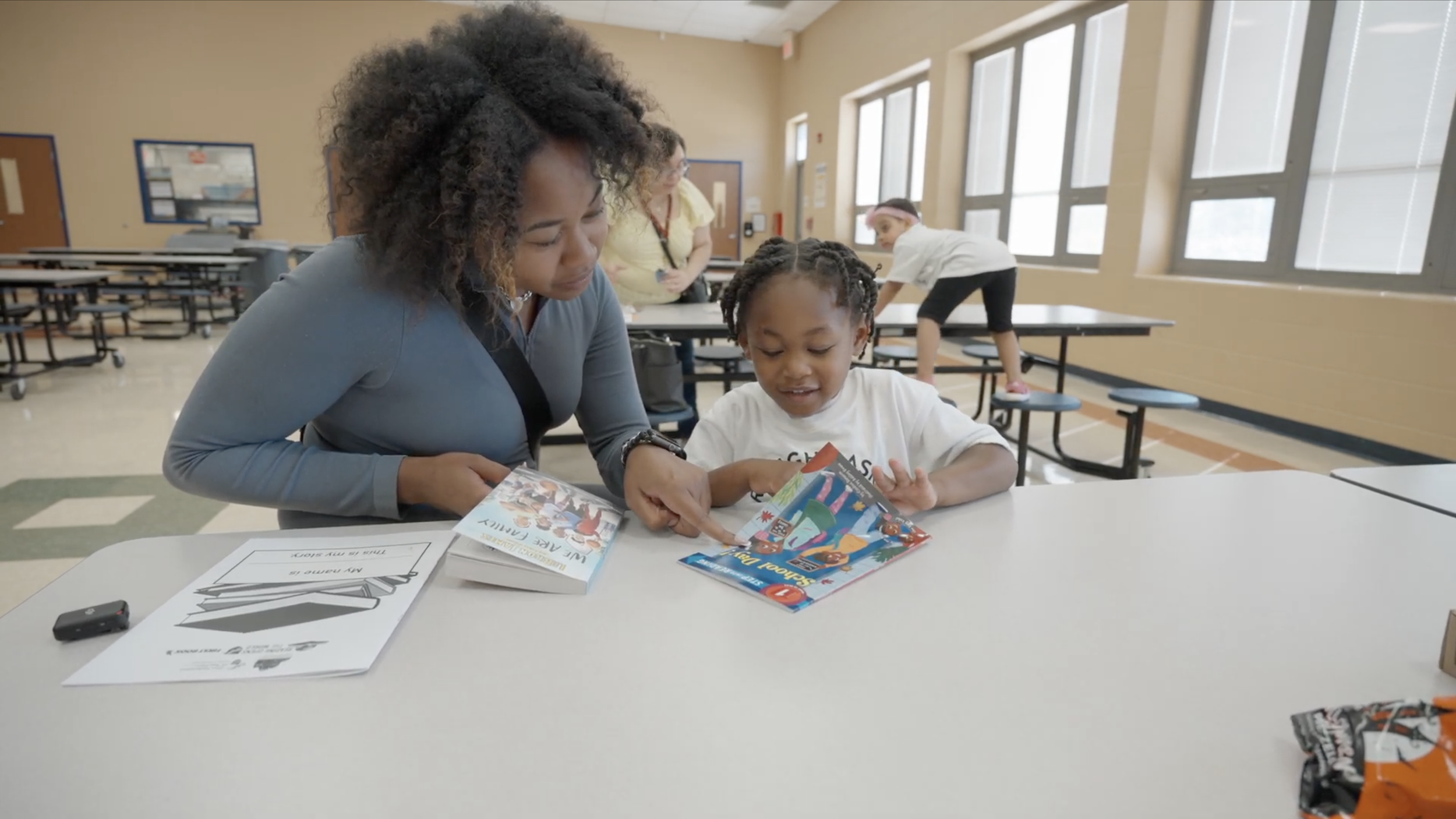 Adult woman and young girl sitting at a school cafeteria table reading colorful, illustrated books, with other children in the background.