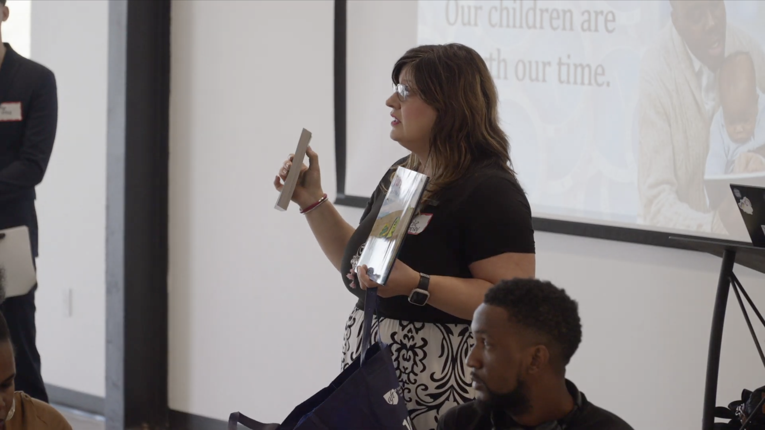A woman with glasses, brown hair, wearing a black shirt and patterned pants, is holding a book and speaking in front of a group during a presentation or seminar. A man with curly hair in the foreground is listening, and a large projector screen is visible in the background.