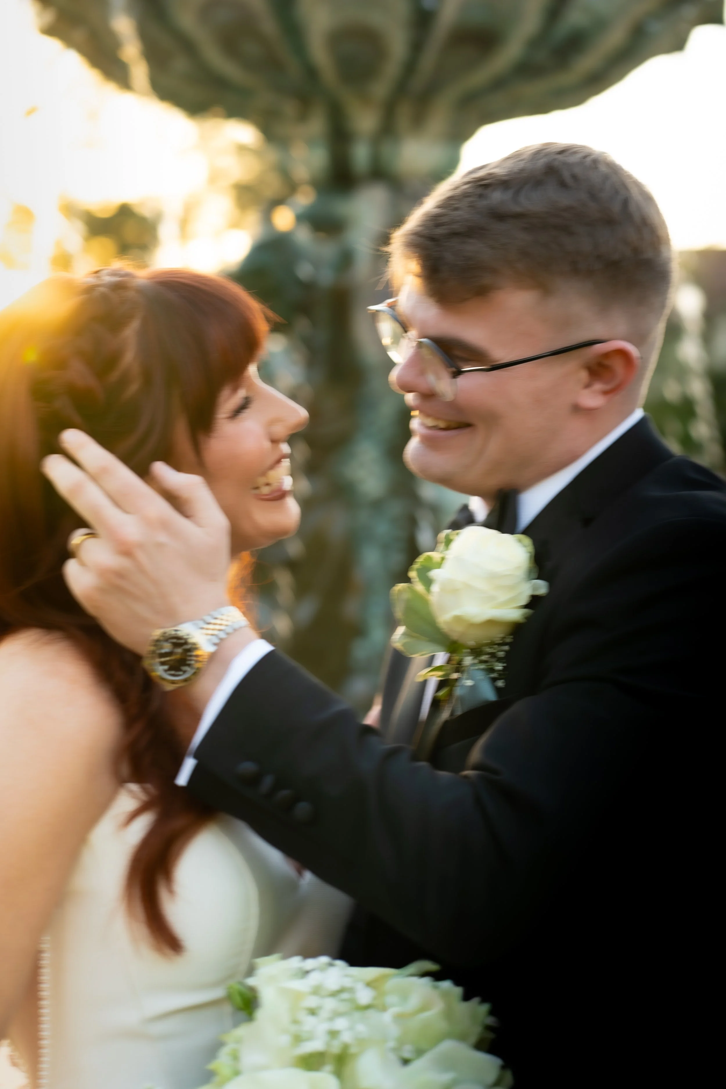 A couple, dressed in wedding attire, is smiling and looking at each other during their wedding ceremony outdoors at sunset. The woman has red hair and is wearing a white dress, while the man wears glasses and a tuxedo with a white rose boutonniere.
