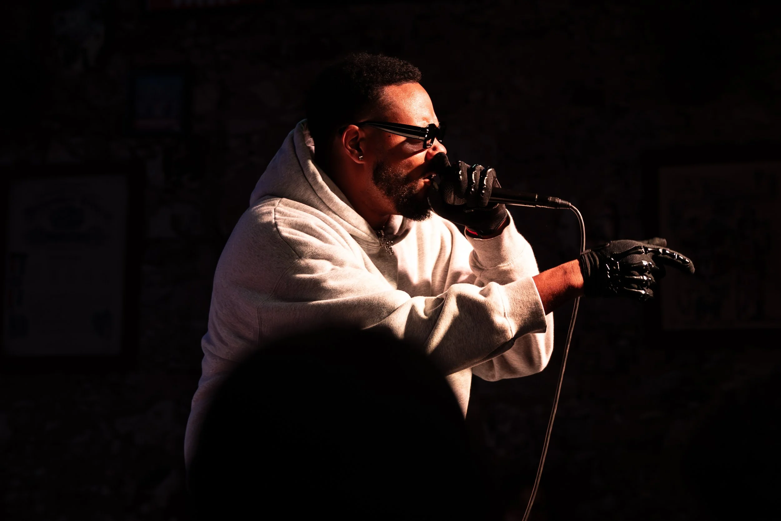 Man wearing glasses and black gloves, sitting in a dark room with warm light, speaking into a microphone and wearing a gray hoodie.