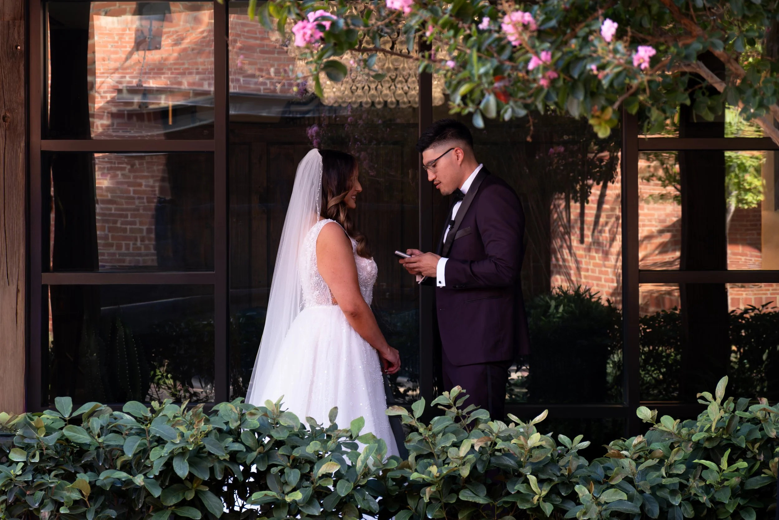 A bride and groom standing face to face outdoors under a flowering tree, with a background of a brick building and large window reflection.