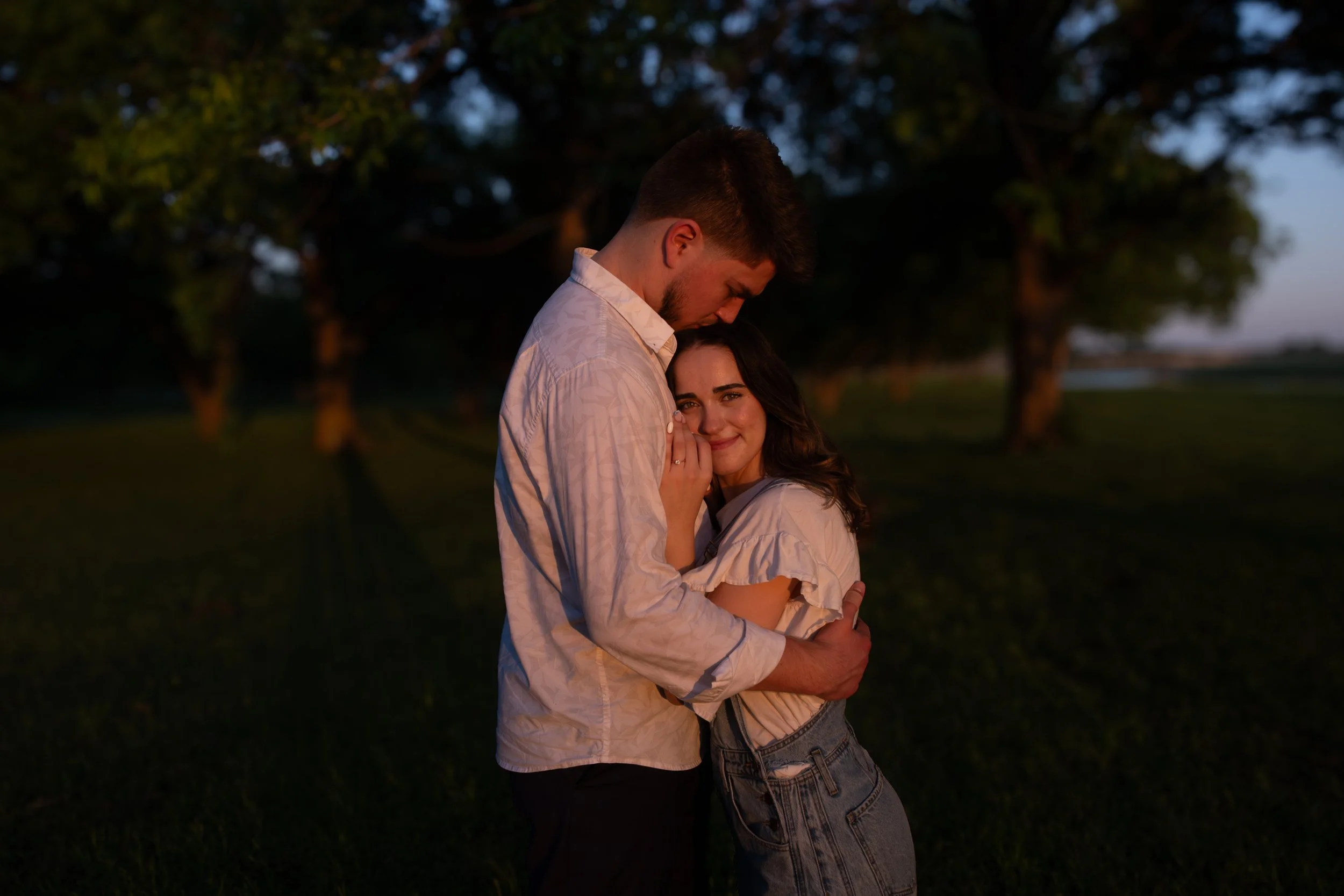 A young couple embracing outdoors at sunset, with trees in the background.